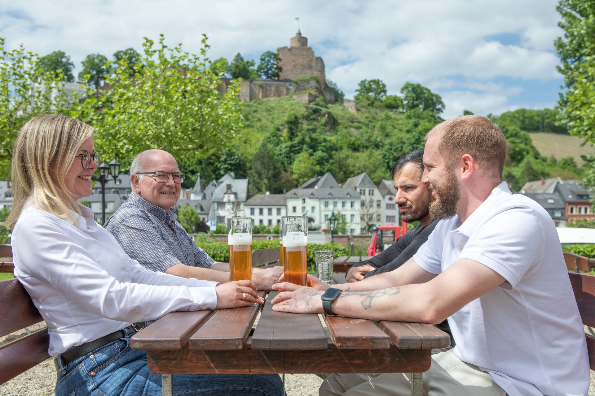 Vier Personen an einem Tisch mit Bier, lächelnd. Fluss und Schloss im Hintergrund an einem sonnigen Tag.