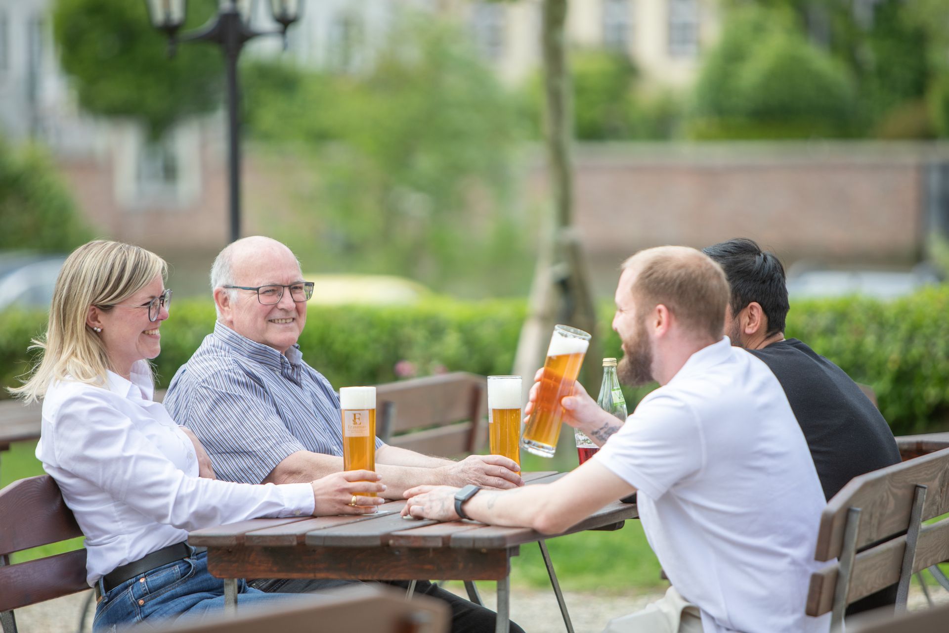Vier Personen sitzen im Freien, stoßen mit Bier an und lächeln sich an.