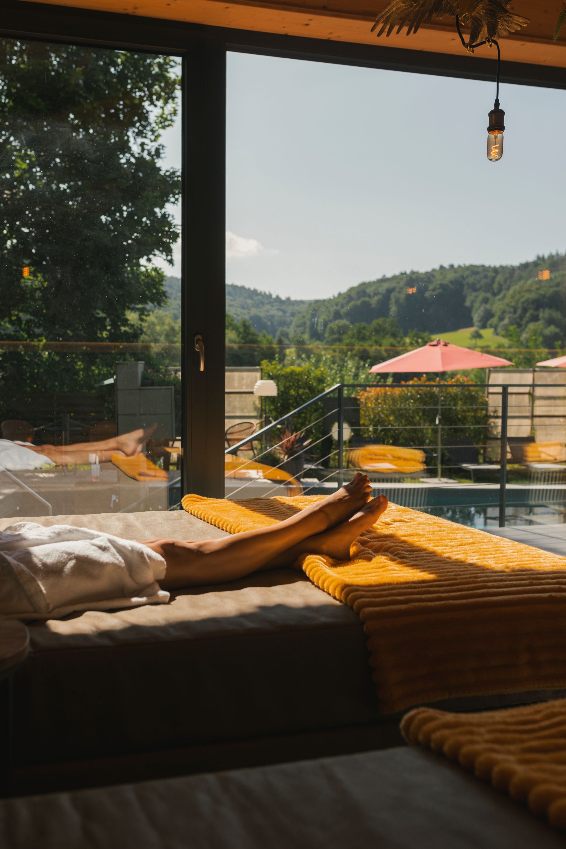 Frau entspannt sich auf einer gelb gedeckten Liege und blickt auf eine Landschaft mit Hügeln, Pool und rotem Dach.