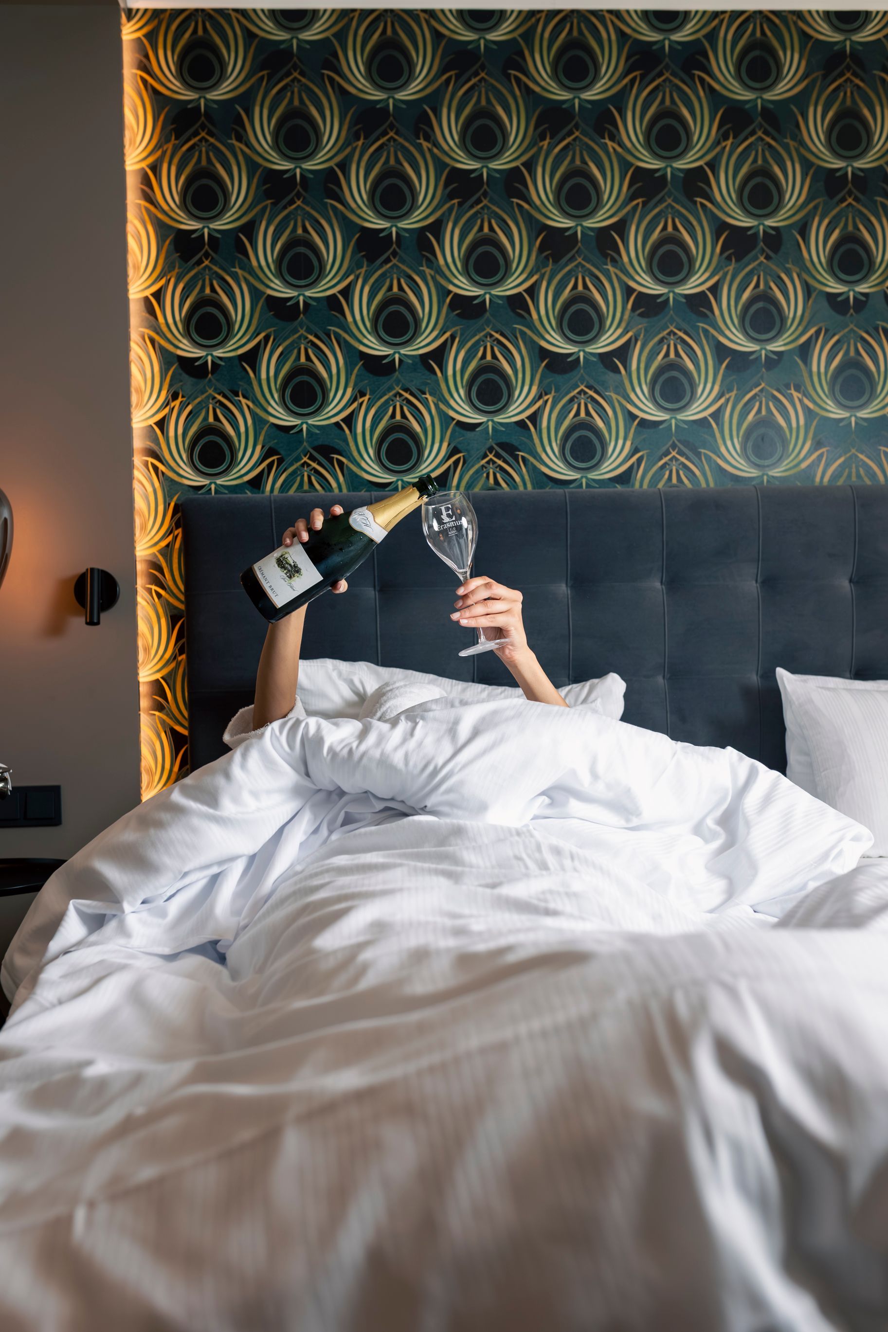 Person im Bett, die Champagner in ein Glas gießt, vor einer Wand mit Pfauenfedermuster.