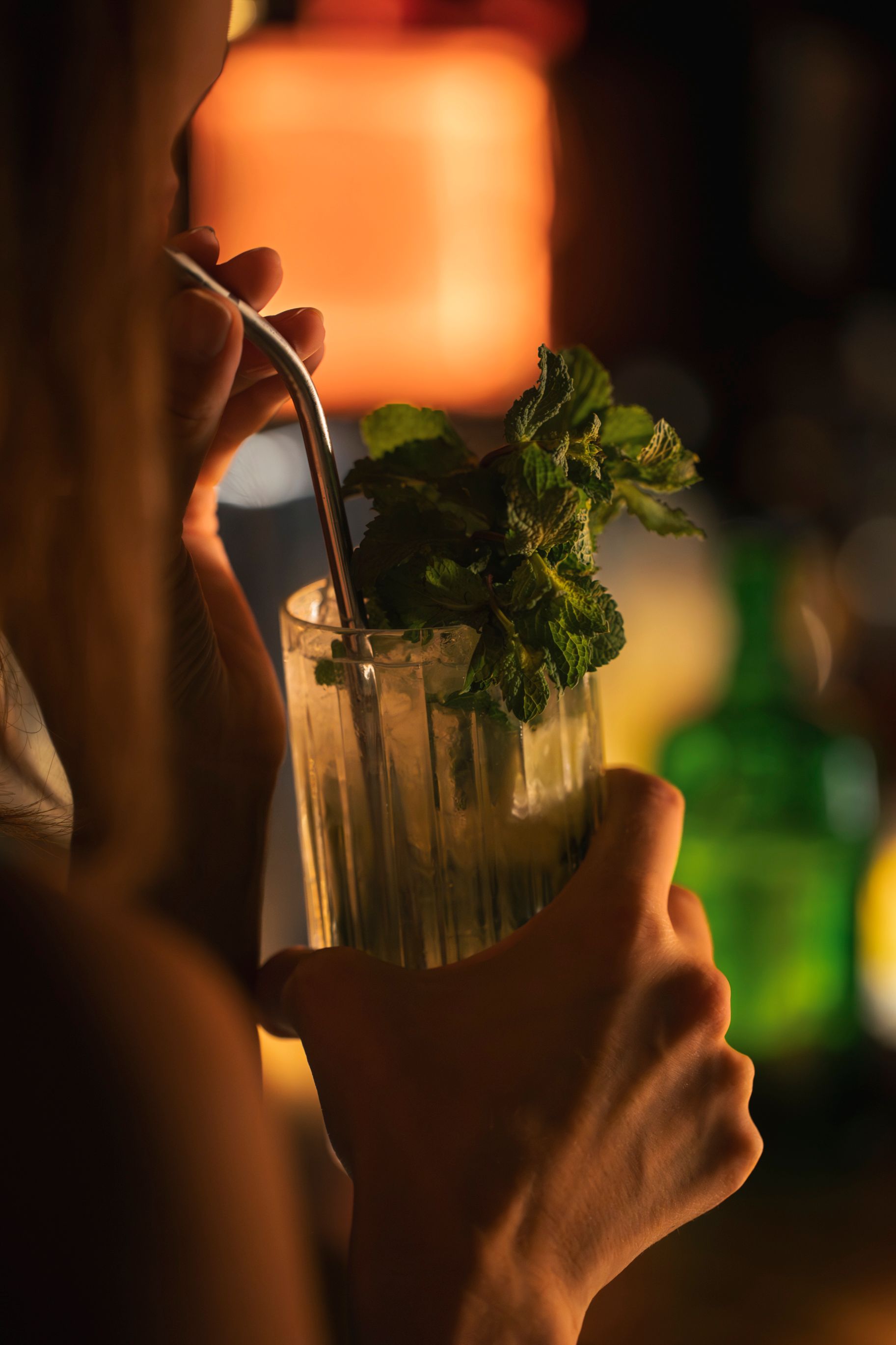 Person hält ein Glas mit einem Cocktail, Minzgarnitur und einem Metallstrohhalm in einer schwach beleuchteten Bar.
