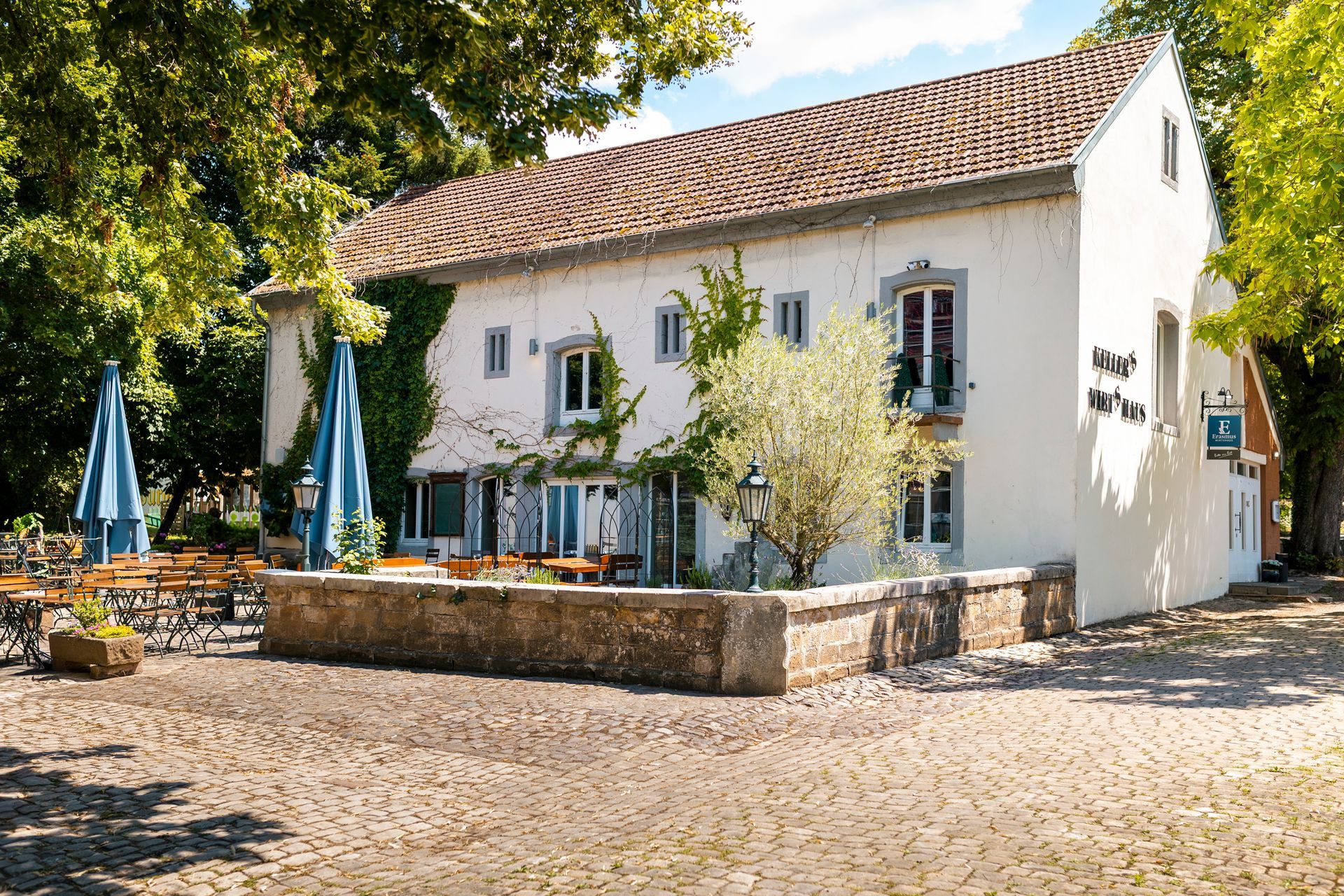 Weißes Gebäude mit braunem Ziegeldach, Sitzgelegenheiten im Freien. Blaue Sonnenschirme, Bäume.