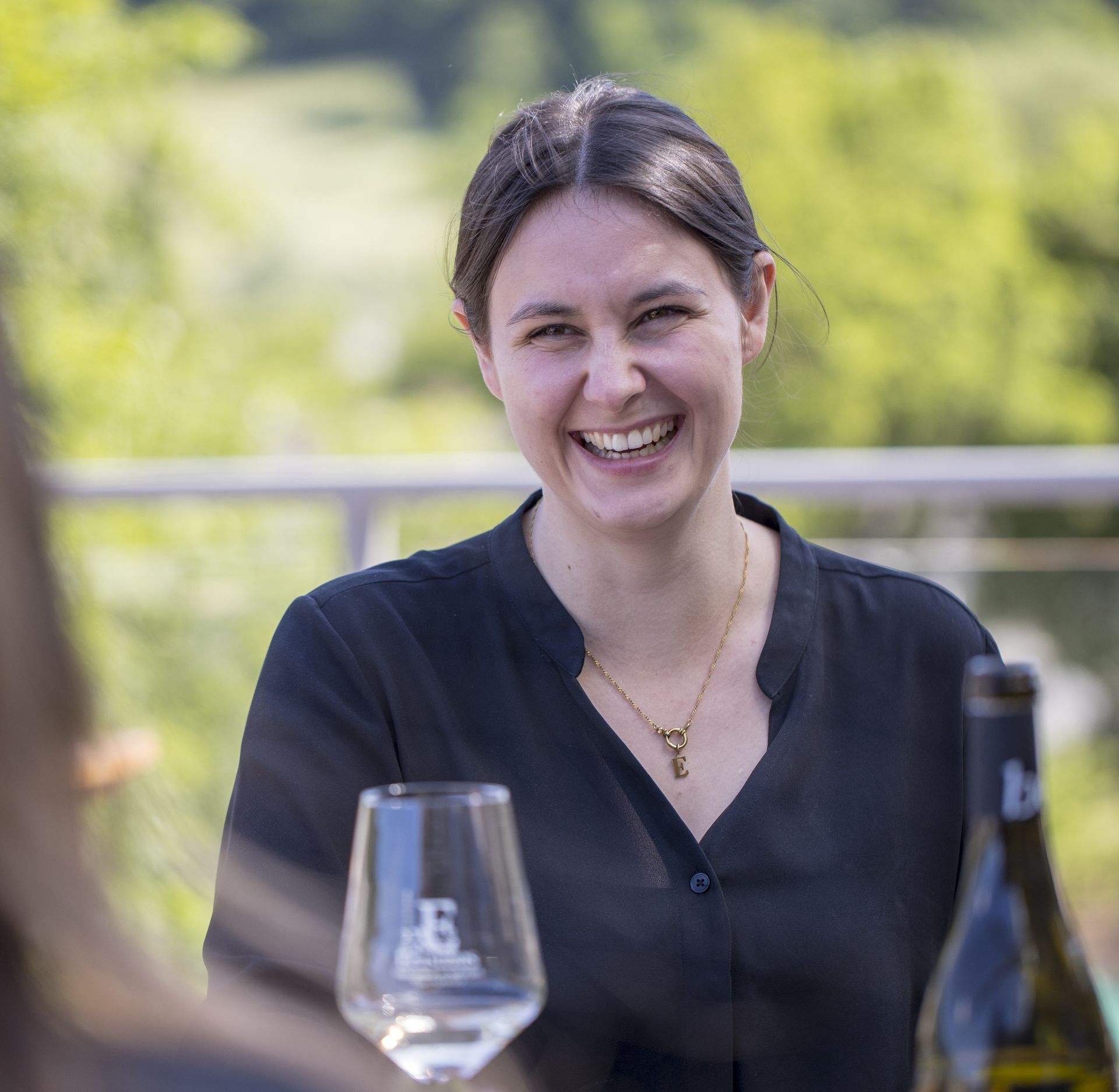 Lächelnde Frau im Freien, neben einem Tisch mit einem Weinglas und einer Flasche. Trägt schwarzes Hemd, Sonnenlicht.