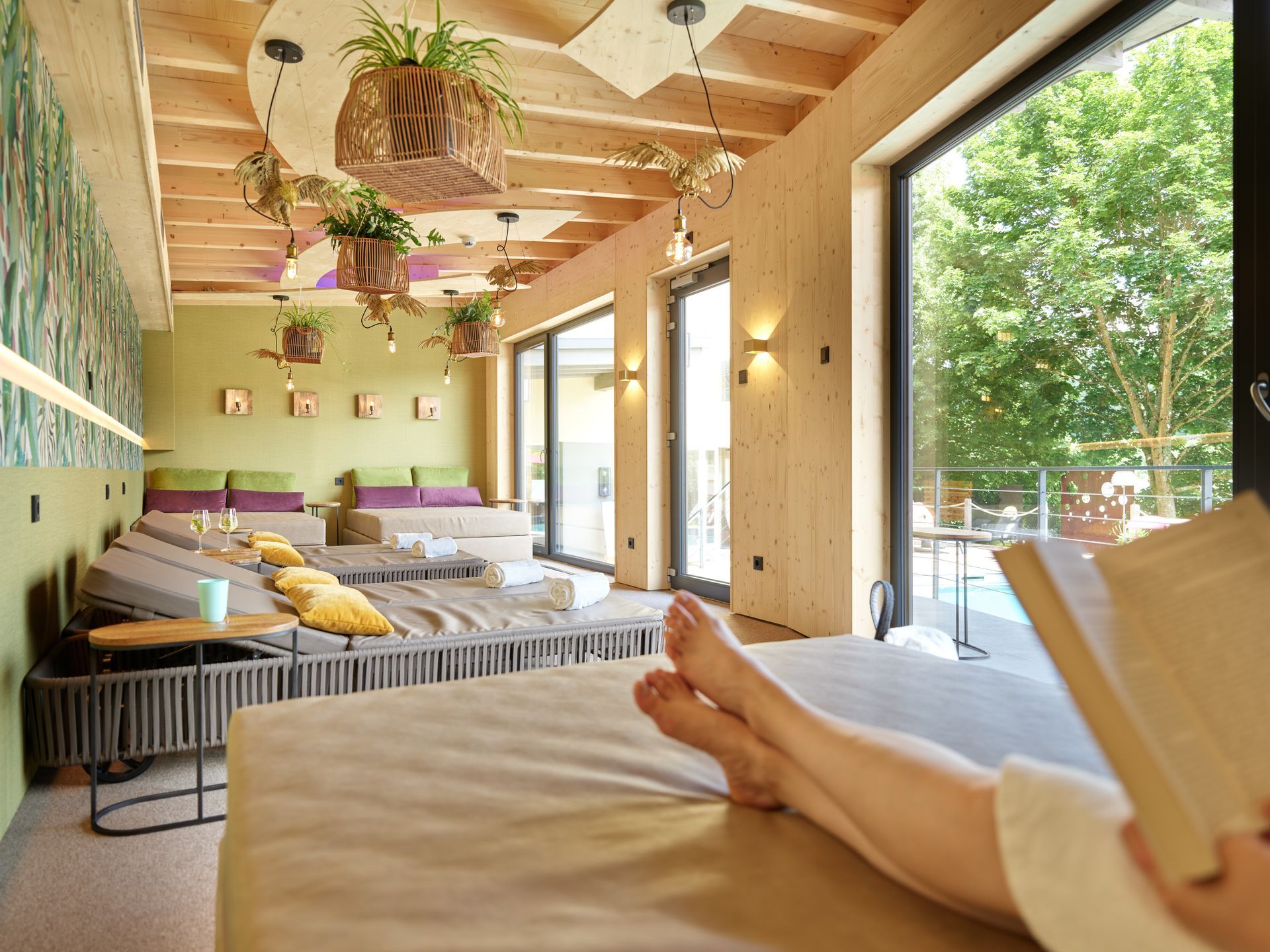 Frau entspannt sich auf einem Liegestuhl in einem Spa und liest ein Buch; im Hintergrund Pflanzen und Fenster.