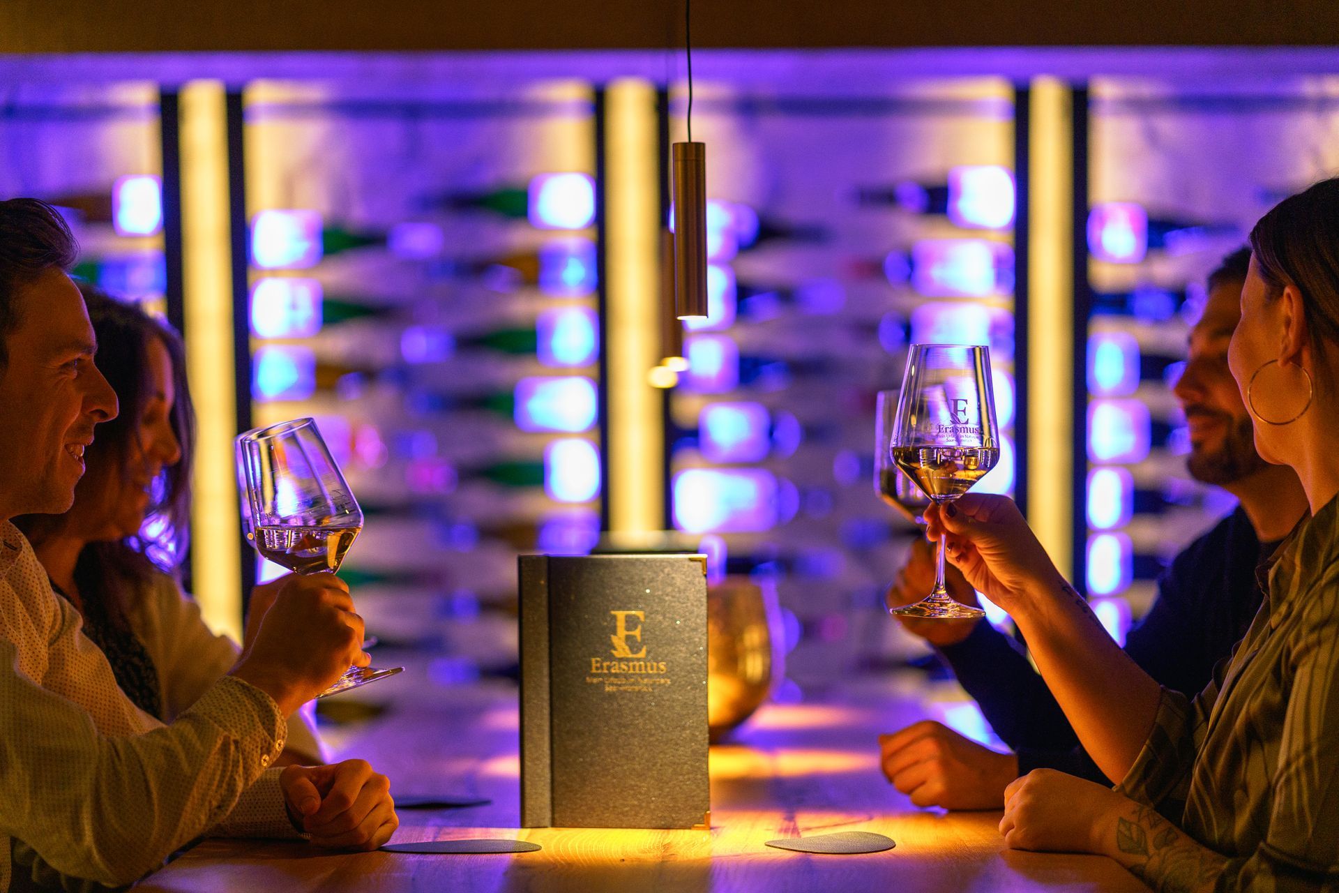 Menschen stoßen mit Weingläsern an einer Bar mit Speisekarte, Flaschen und violetter Beleuchtung an.