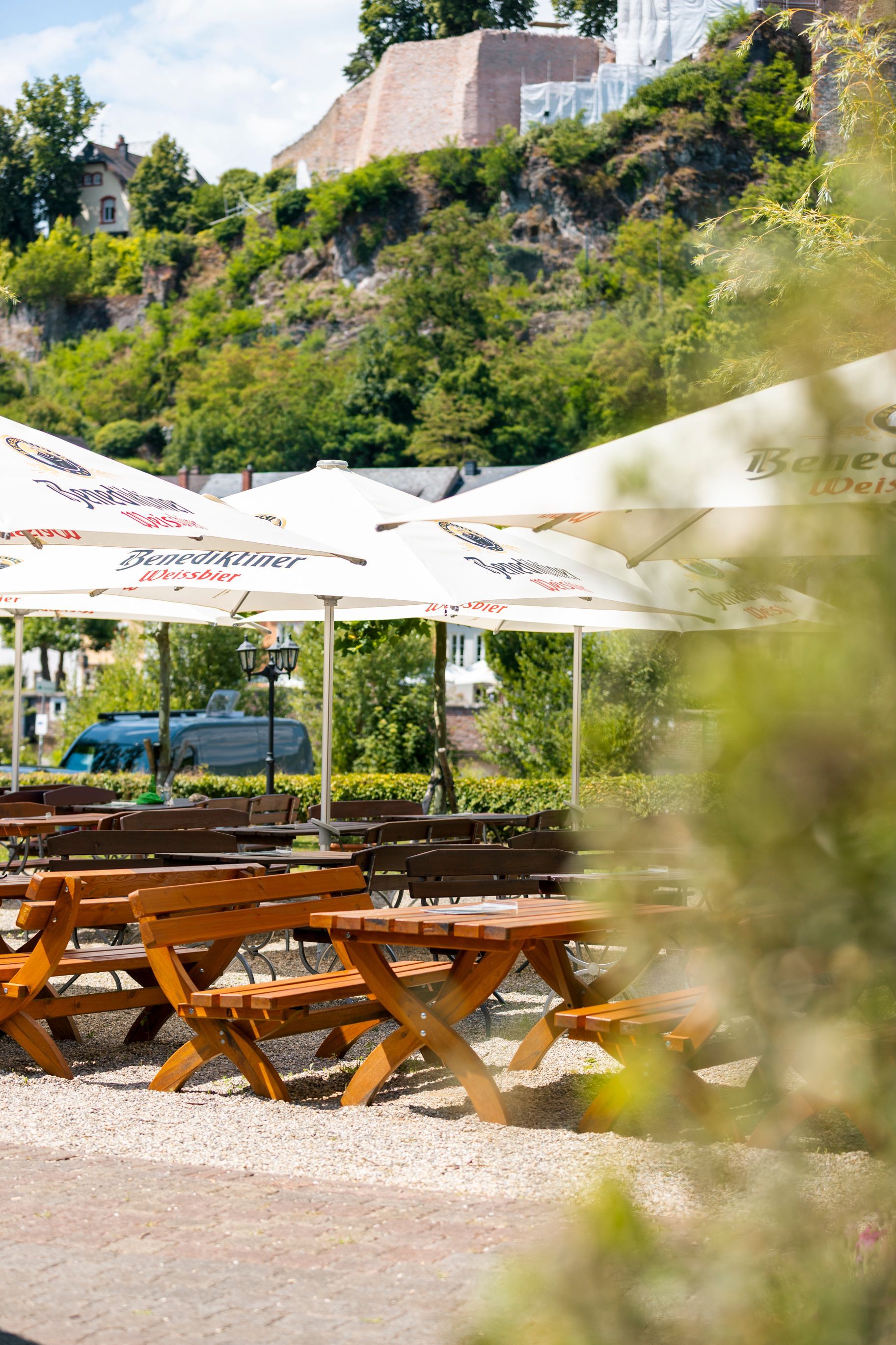 Biergarten im Freien mit Holztischen unter Sonnenschirmen, im Hintergrund ein Hügel.