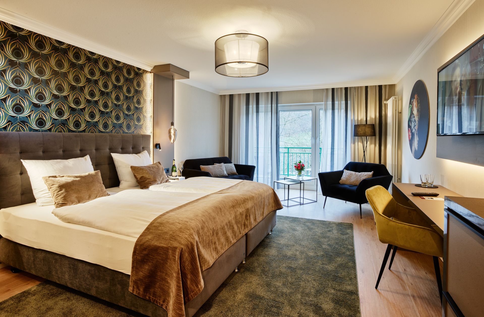 Hotelzimmer mit Kingsize-Bett, Sitzecke und Schreibtisch. Mit gemusterter Tapete und Fenster.