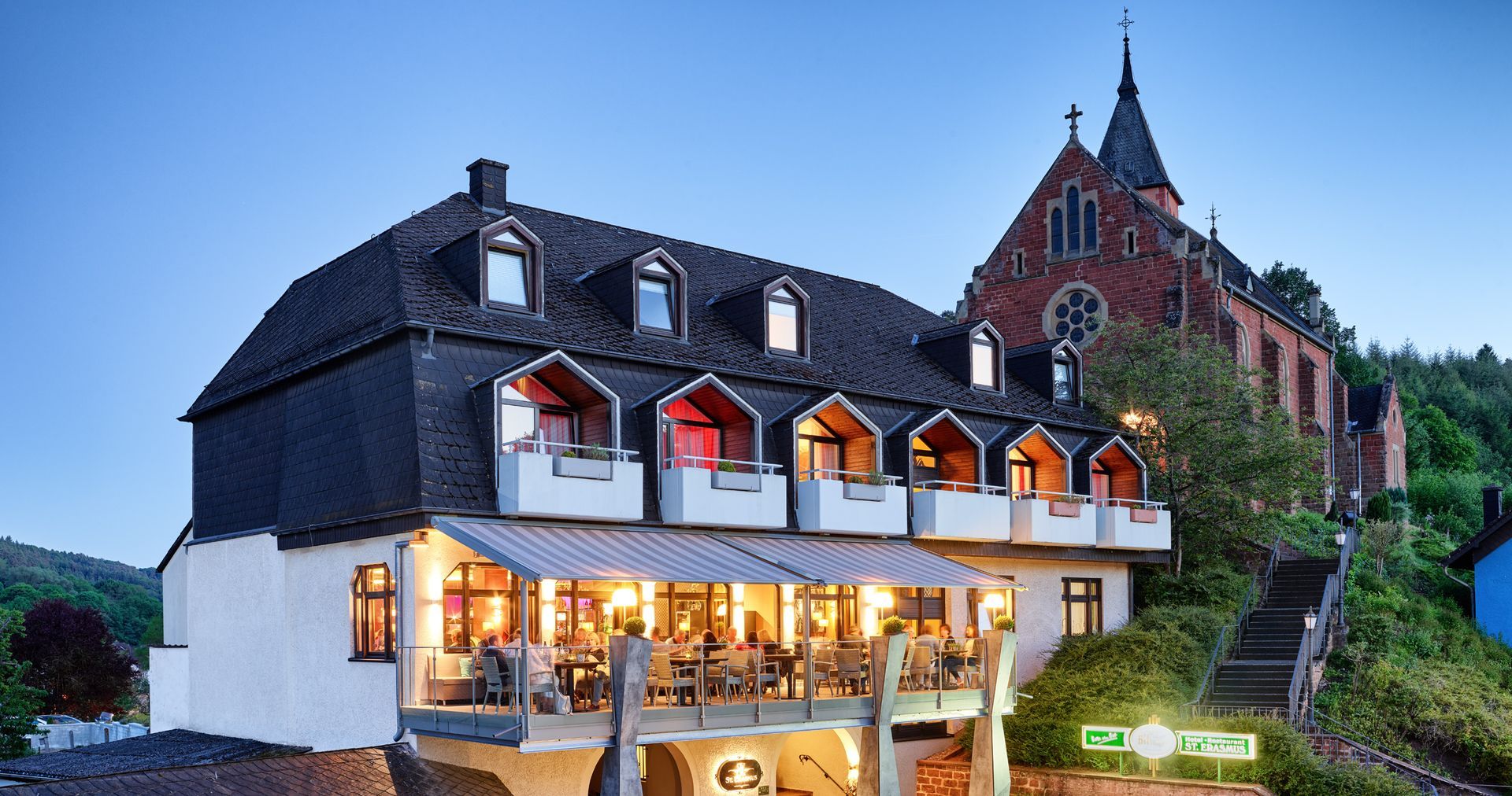 Hotel mit Sitzgelegenheiten im Freien, dunklem Dach, neben einer Kirche auf einem Hügel in der Abenddämmerung.