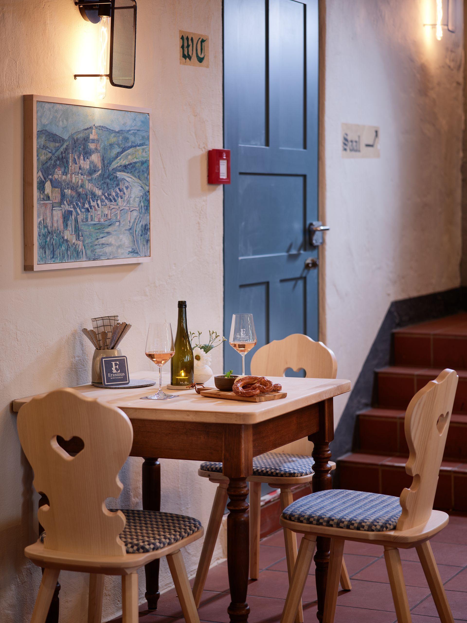Gemütliche Restaurantecke: Holztisch für zwei Personen, Stühle mit Rückenlehne, blaue Tür, kleines Gemälde und Treppe.