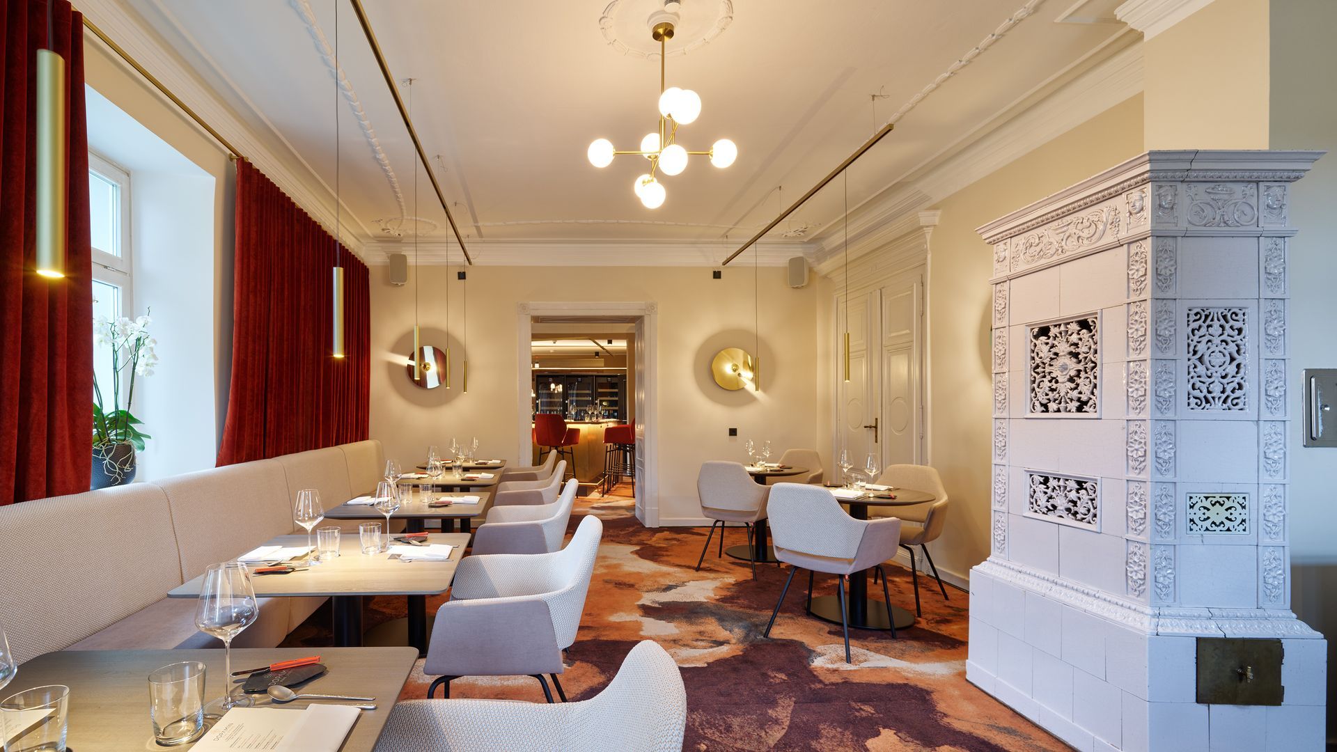 Elegantes Restaurant-Interieur mit Tischen, Stühlen, roten Vorhängen und dekorativem weißen Ofen.