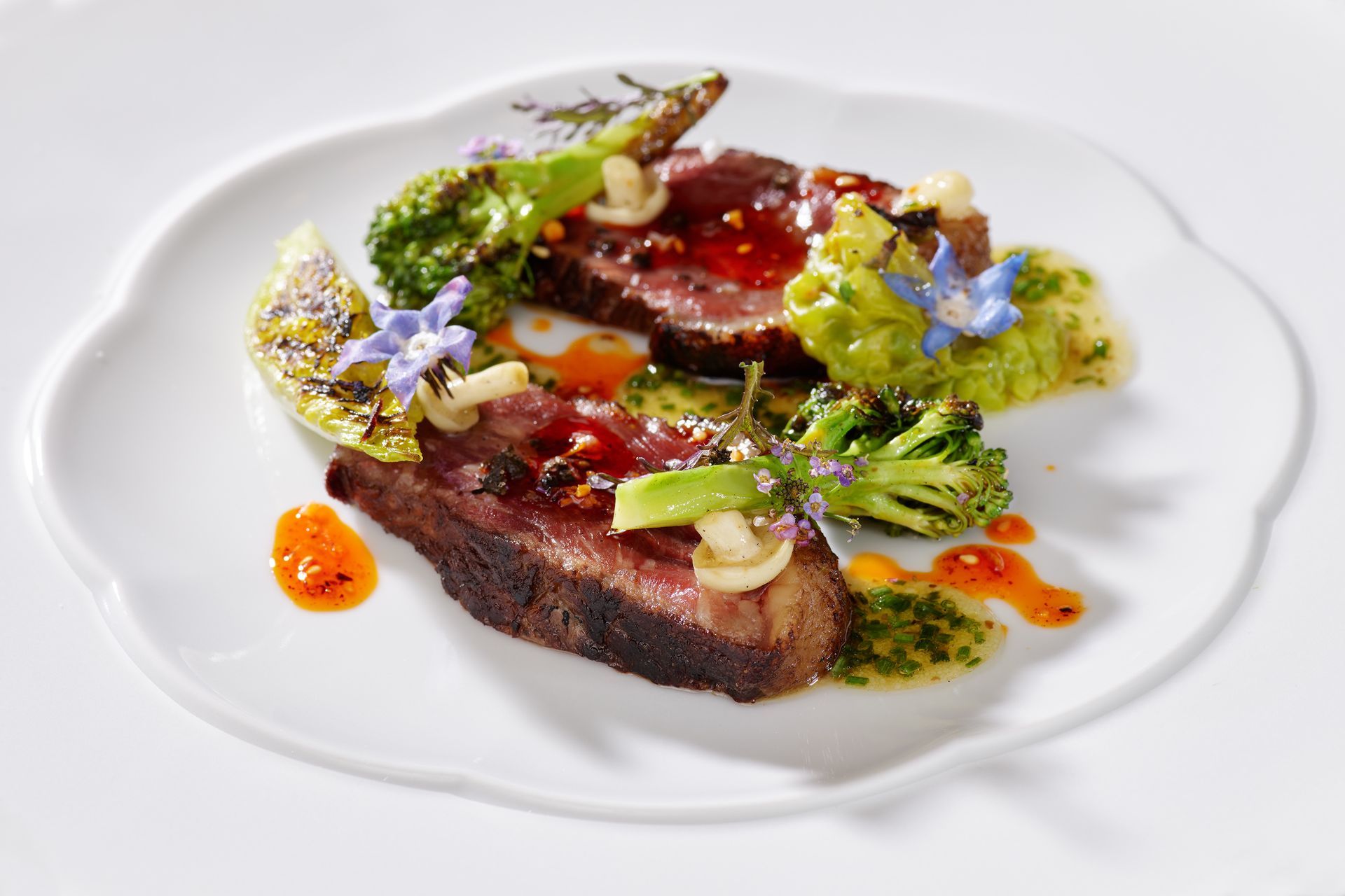 Gebratene Rindfleischscheiben mit geröstetem Brokkoli, Saucentropfen und essbaren Blüten auf einem weißen Teller.