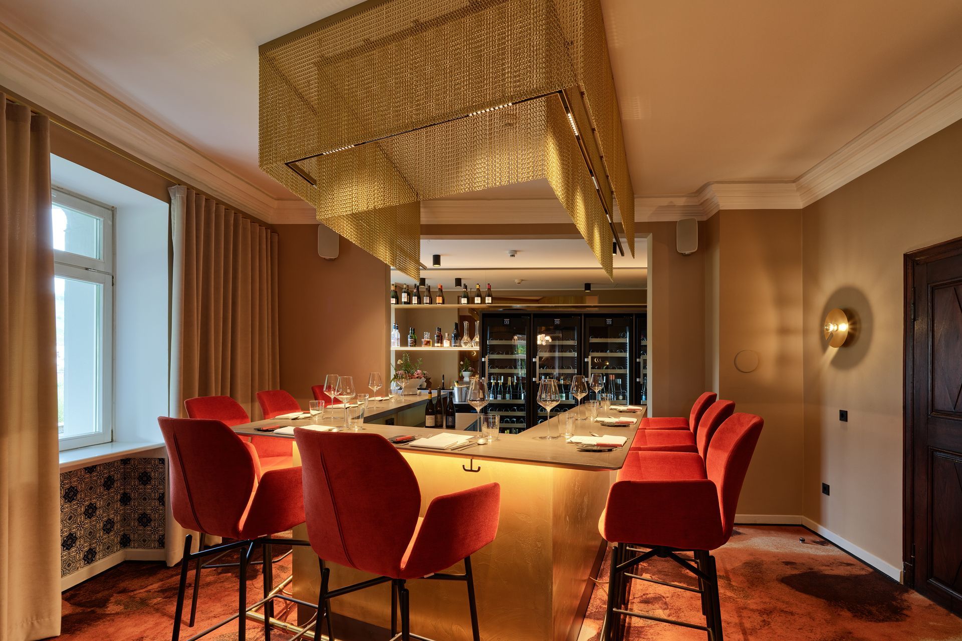 Eine Bar mit roten Hockern, goldenem Kronleuchter und einer Weinauswahl in einem warm beleuchteten Raum.