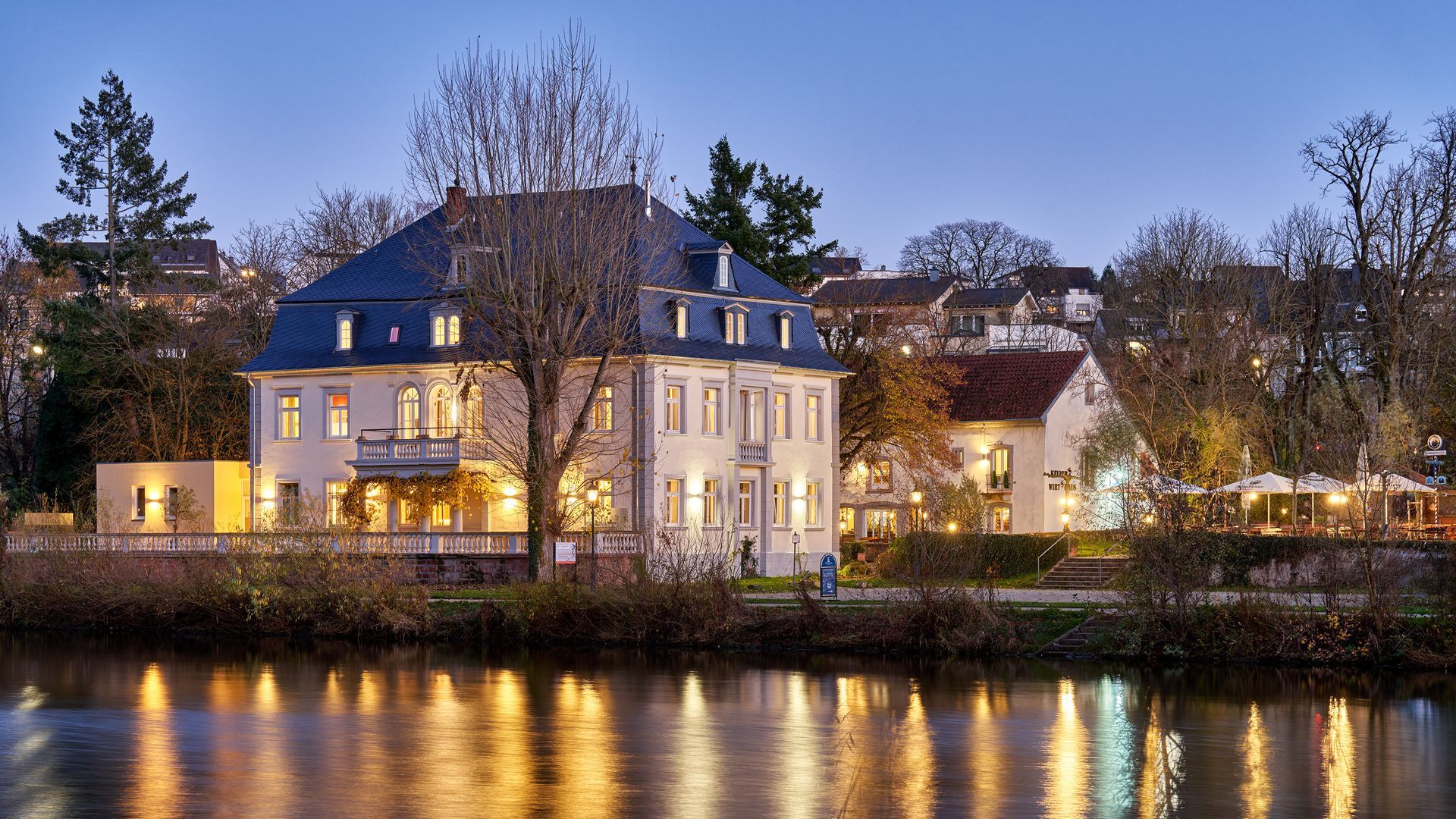 Ein Gebäude am Flussufer, in der Abenddämmerung, spiegelt sich im Wasser. Es sind weitere Gebäude und Bäume zu sehen.