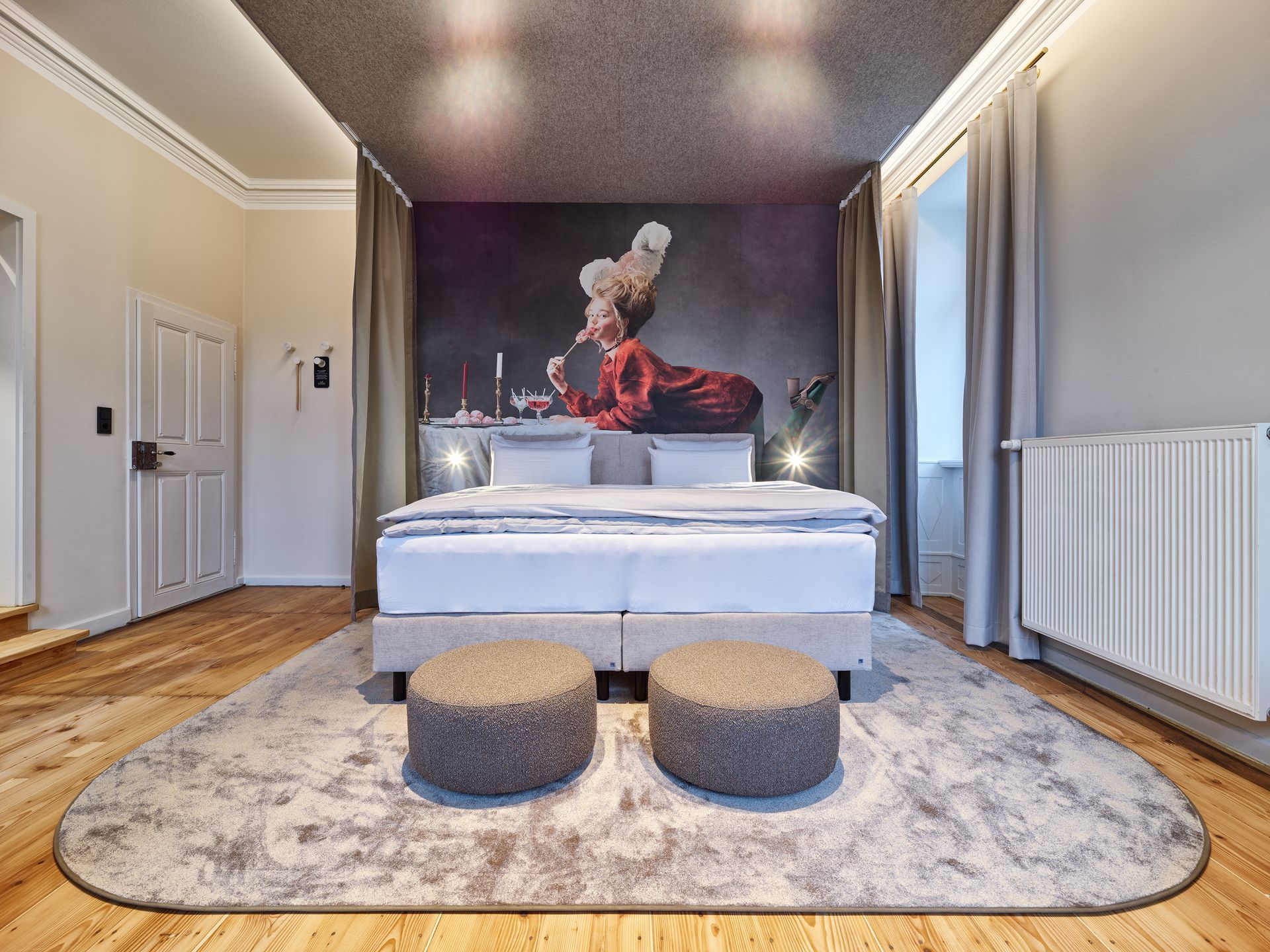 Schlafzimmer mit großem Bett, kunstvollem Wandgemälde, zwei runden Hockern und Holzboden.