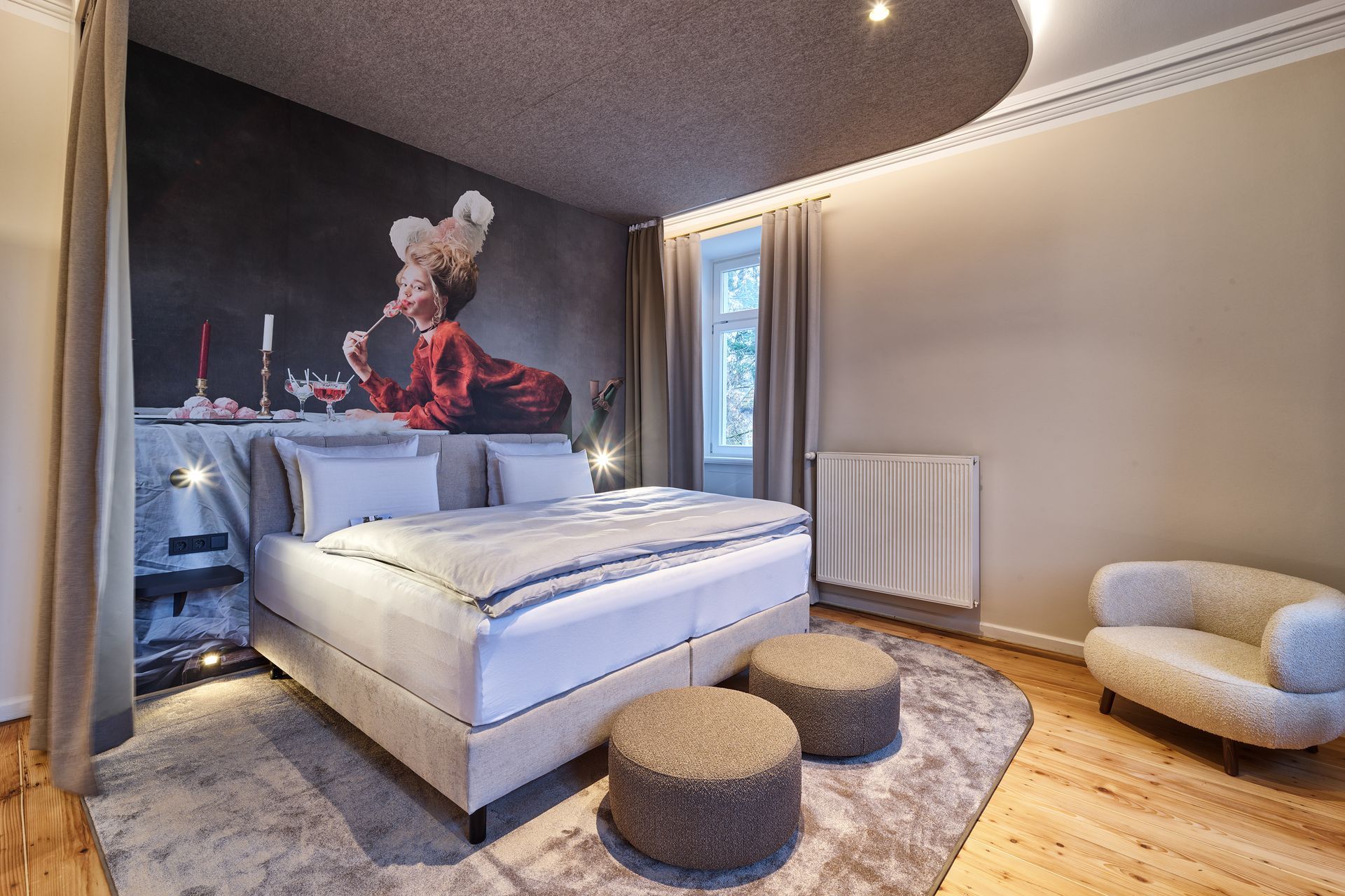 Schlafzimmer mit einem großen Bett, einem kunstvollen Wandgemälde, zwei kleinen Hockern und einem Sessel auf einem Teppich.