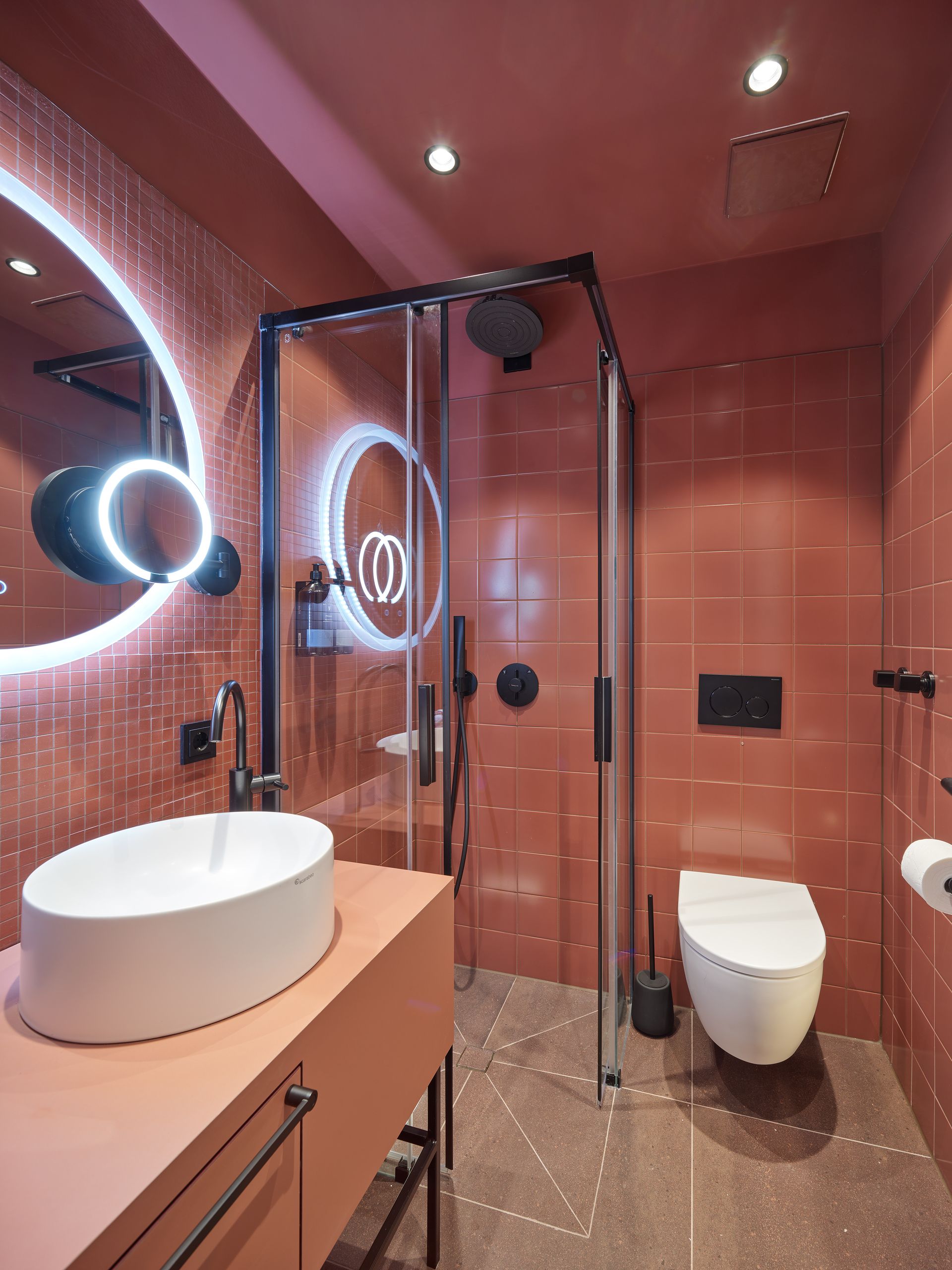 Badezimmer mit roten Fliesen, Glasdusche, moderner Toilette und einem runden Spiegel über einem weißen Waschbecken.