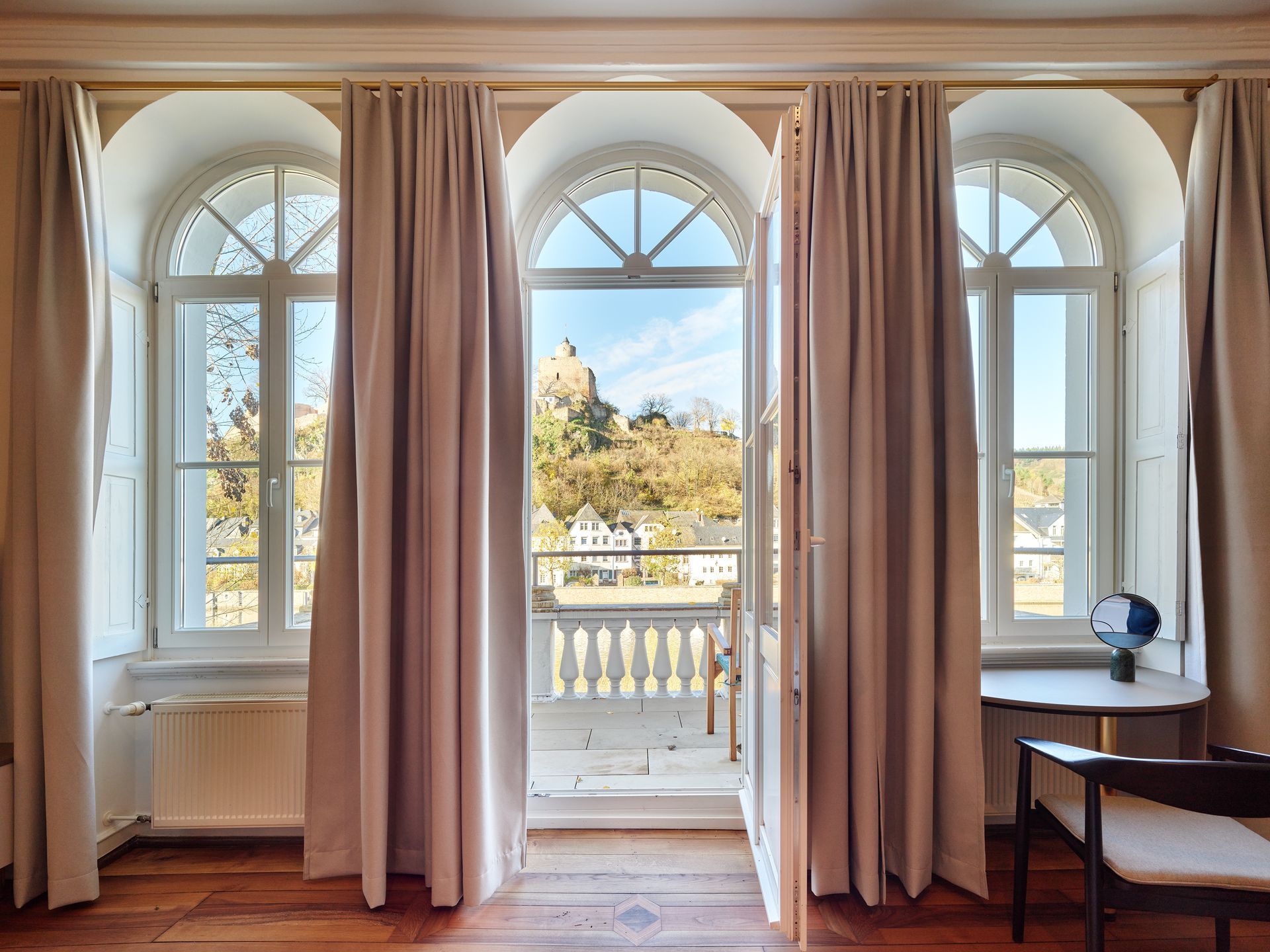 Drei Bogenfenster mit Vorhängen, Balkonblick auf eine Hügelstadt und eine Burg. Holzboden, neutrale Wände.