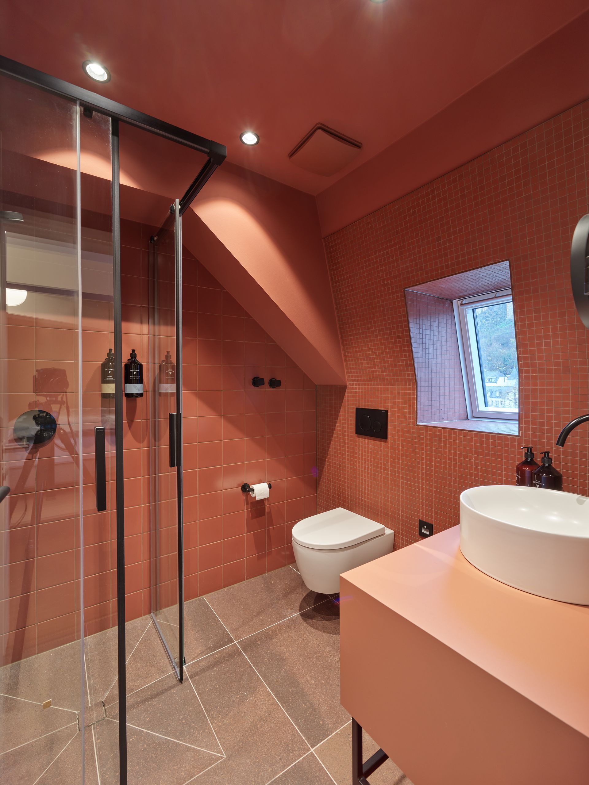 Terrakottafarbenes Badezimmer mit Dusche, WC und Waschbecken. Kleines Fenster, schwarze Armaturen.