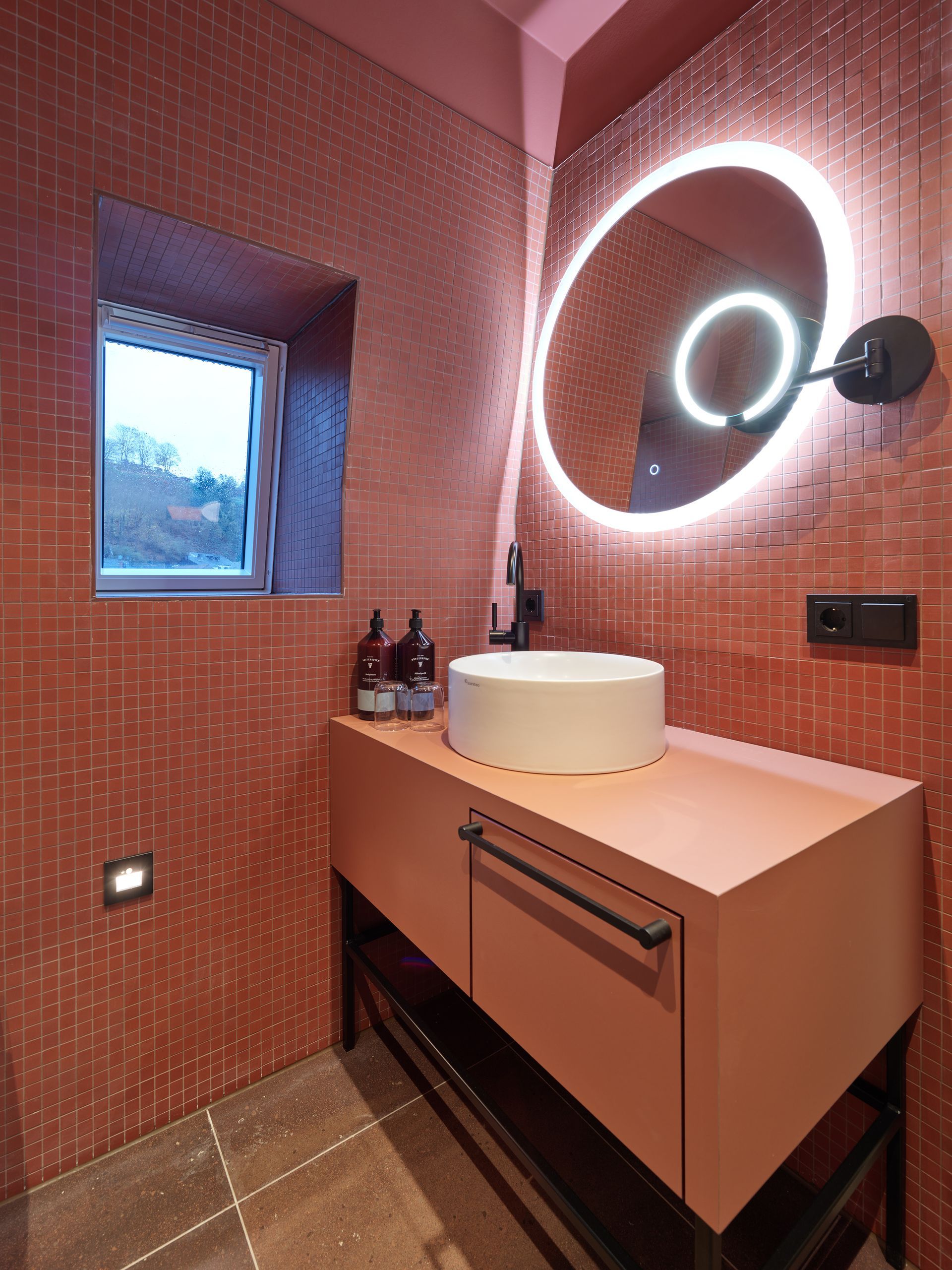 Badezimmer mit roten Fliesen, einem runden Spiegel, einem Waschbecken auf einem roten Schrank und einem kleinen Fenster.