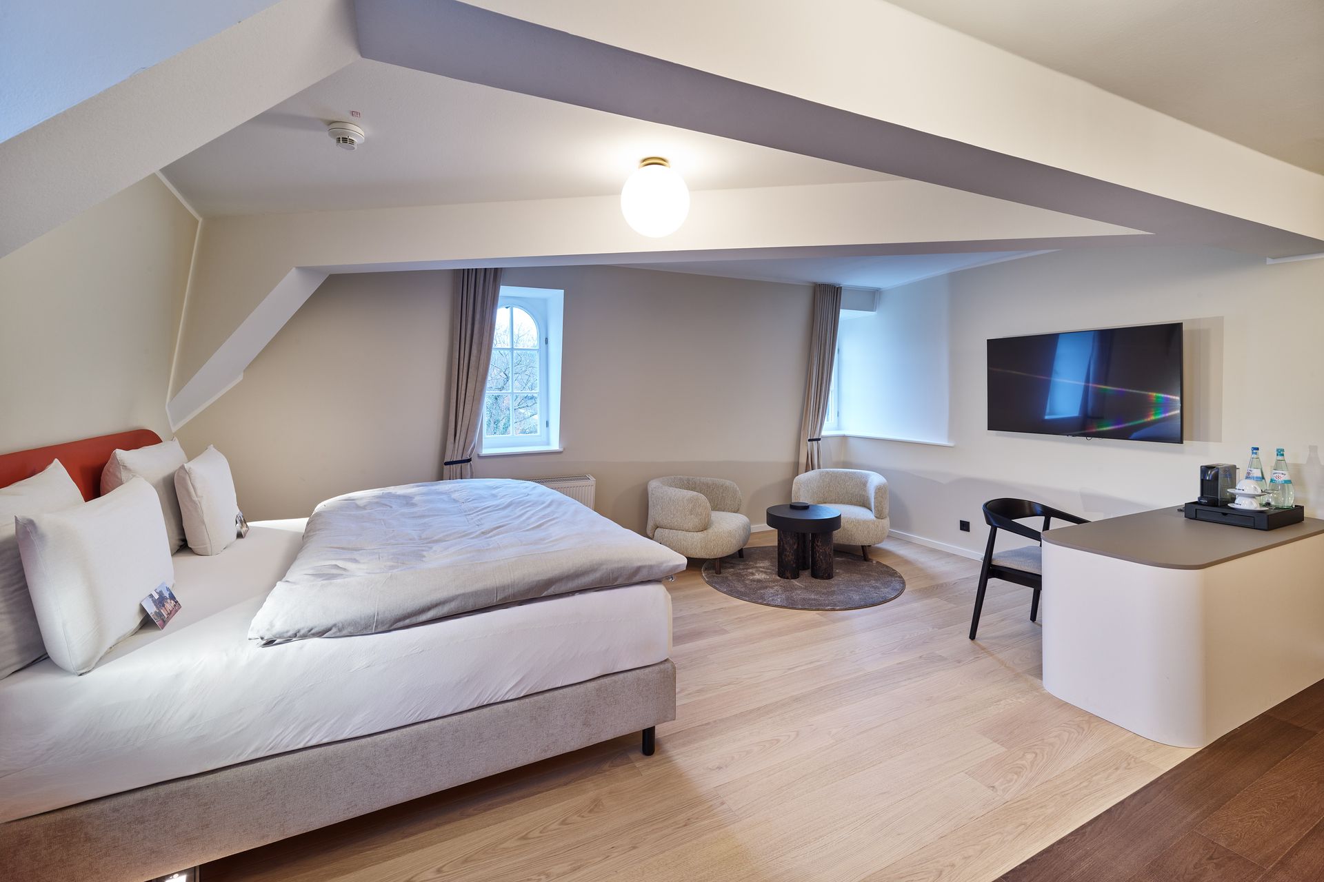 Hotelzimmer mit Bett, Schreibtisch, Stühlen, Fernseher und rundem Teppich.