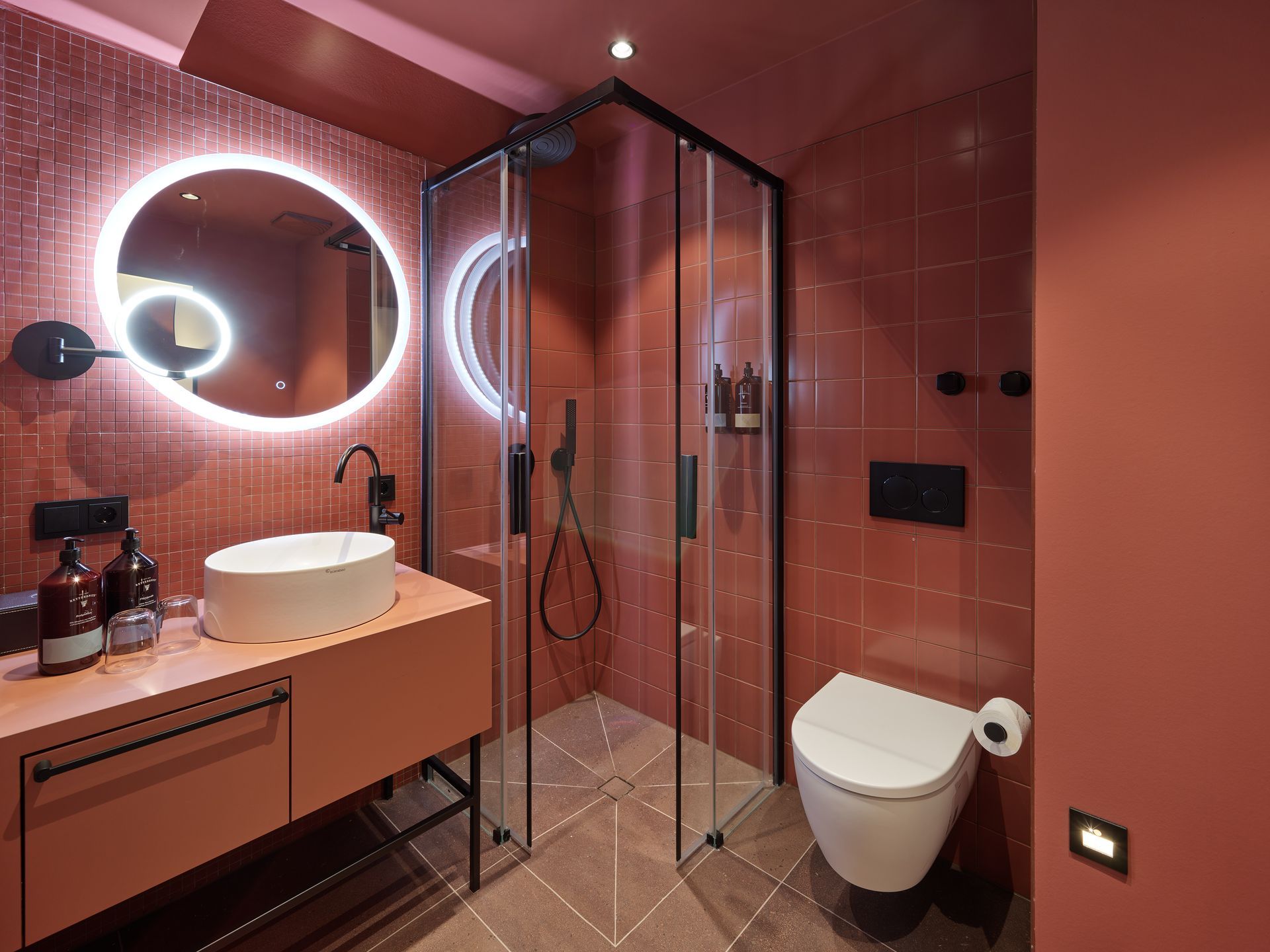 Modernes rosafarbenes Badezimmer mit Glasdusche, rundem Spiegel und schwebendem Waschtisch.