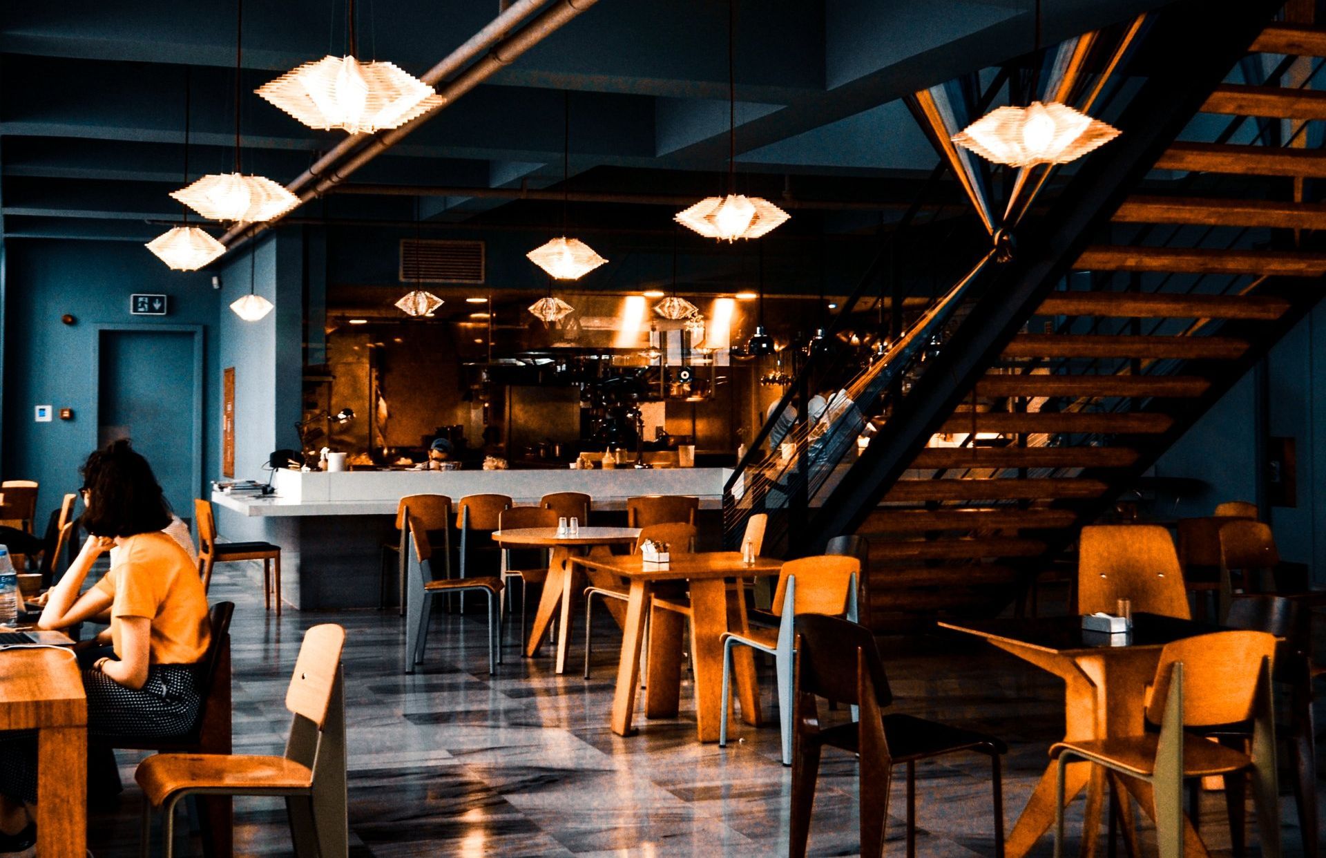 Restaurant interior with tables, chairs, and a staircase. People sit at the tables. Warm lighting.