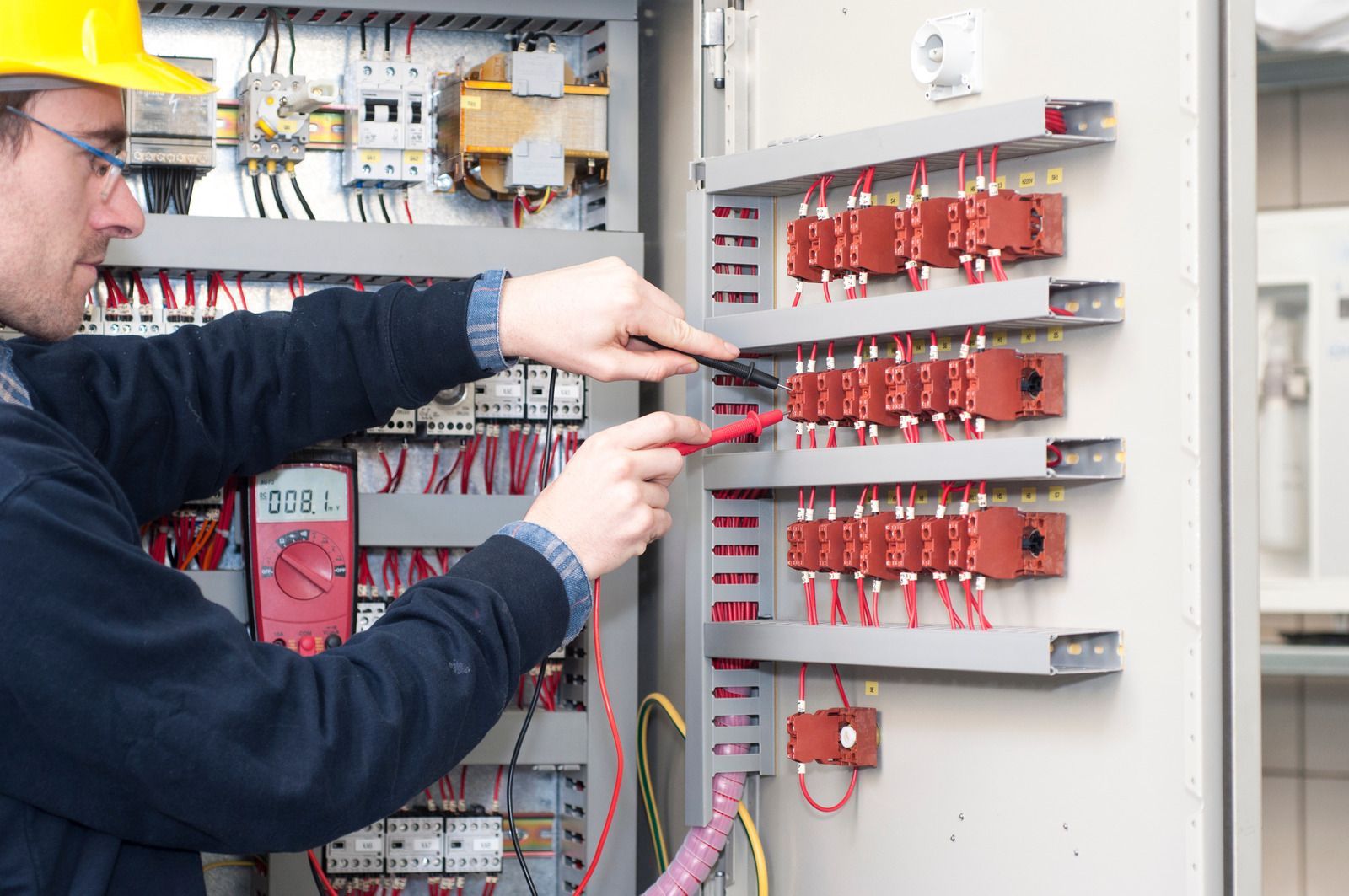Electrician in yellow hard hat using a multimeter to test wires in an electrical panel.