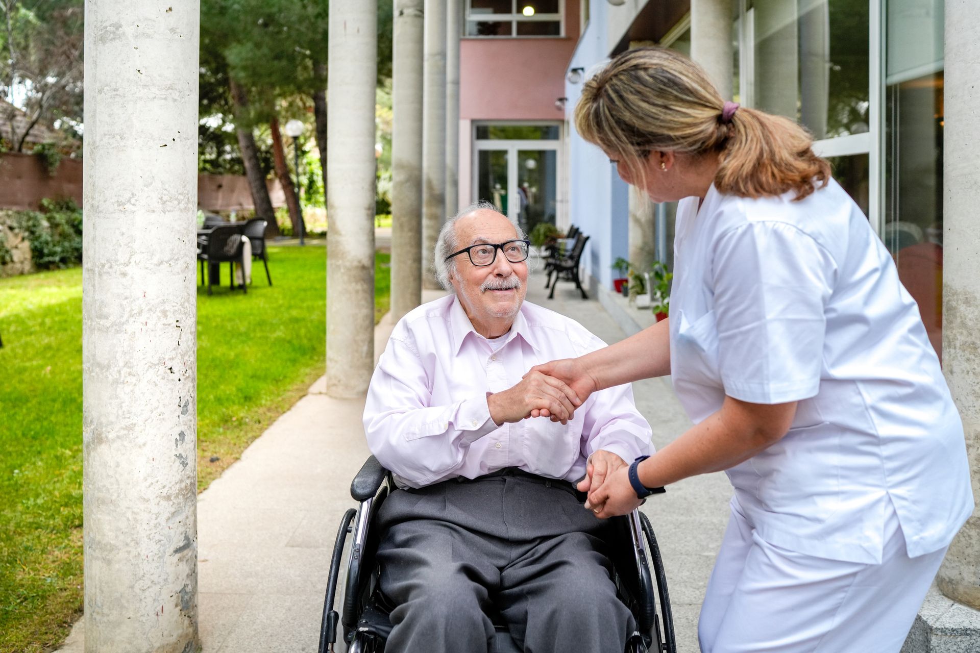 An elderly man in a wheelchair is shaking hands with a nurse.