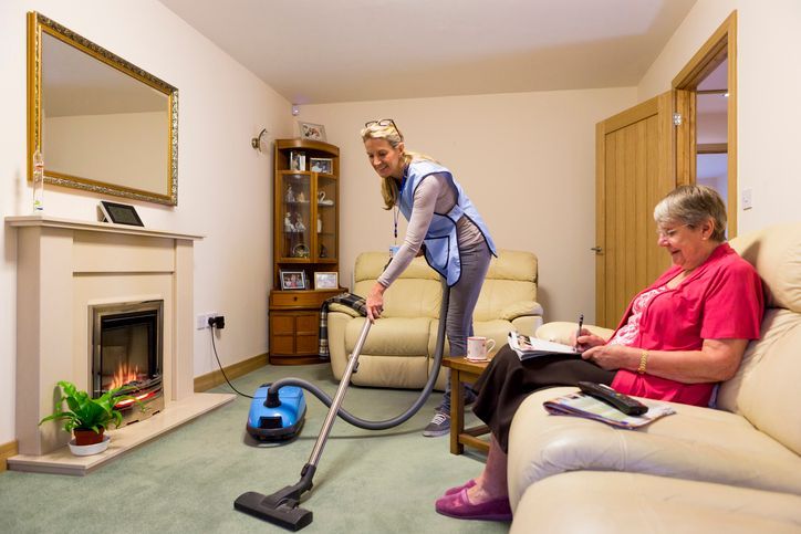 A woman is vacuuming the carpet in a living room while an older woman sits on a couch.