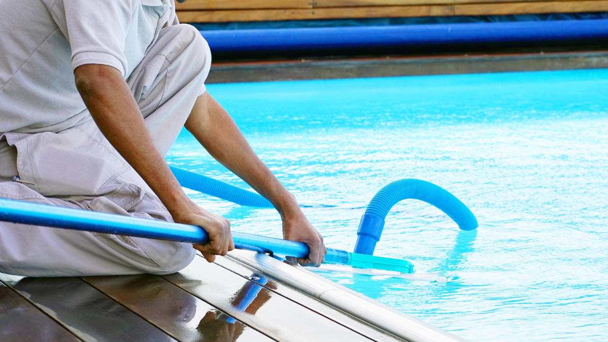Person cleaning a pool with a blue net and pole, next to blue water.