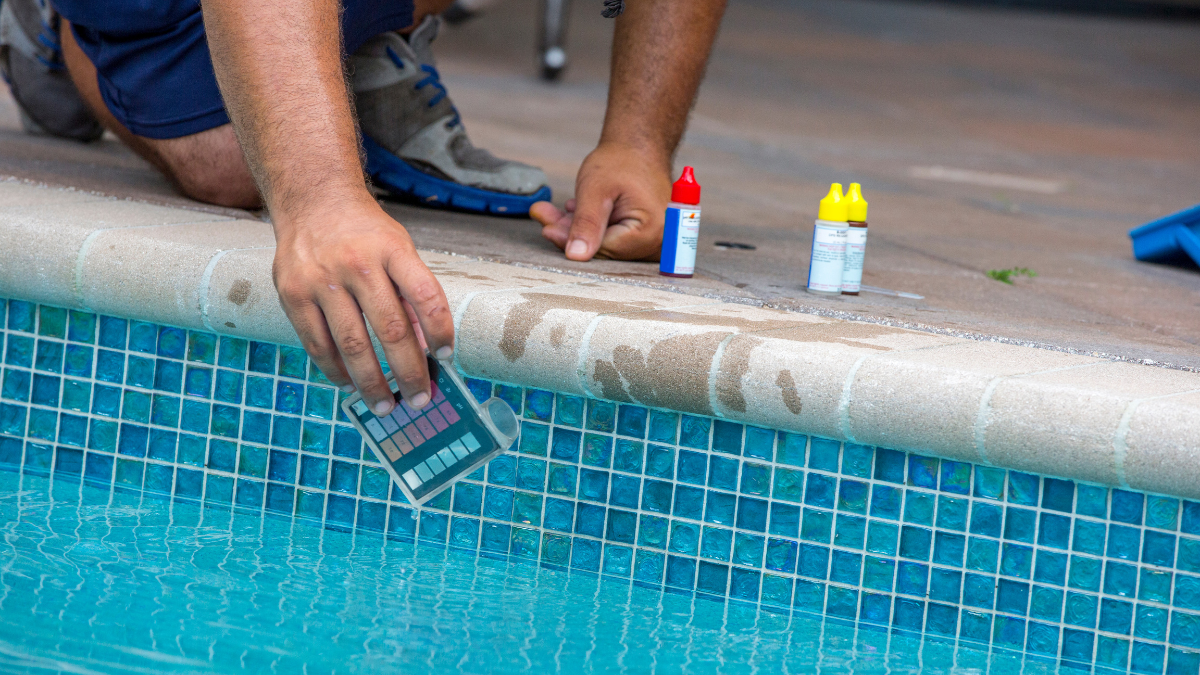 Person testing pool water with test kit and bottles, near a blue tiled pool.