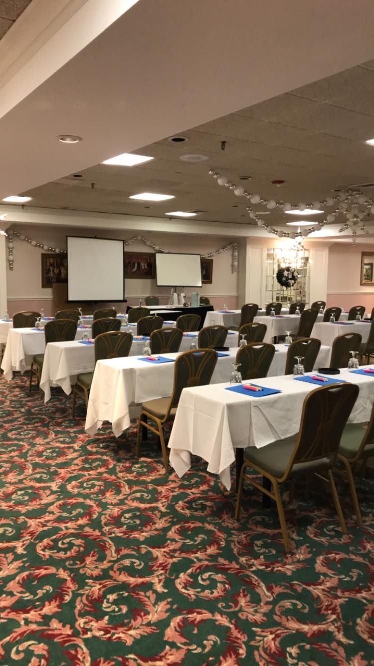 Conference room with tables covered in white tablecloths, arranged for a presentation. Brown chairs and patterned carpet.