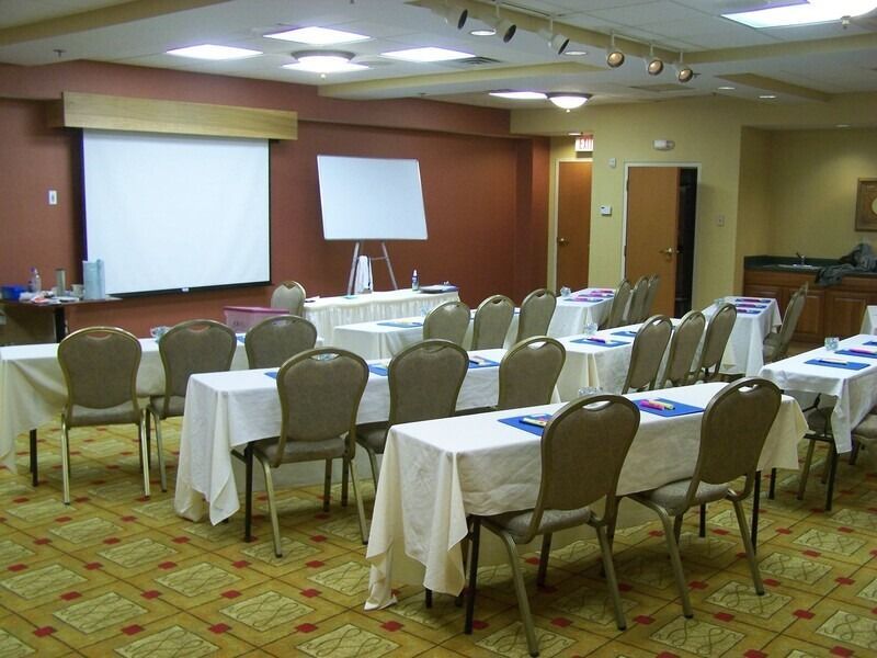 Conference room with tables, chairs, projector screen, and flip chart. Beige and brown tones.