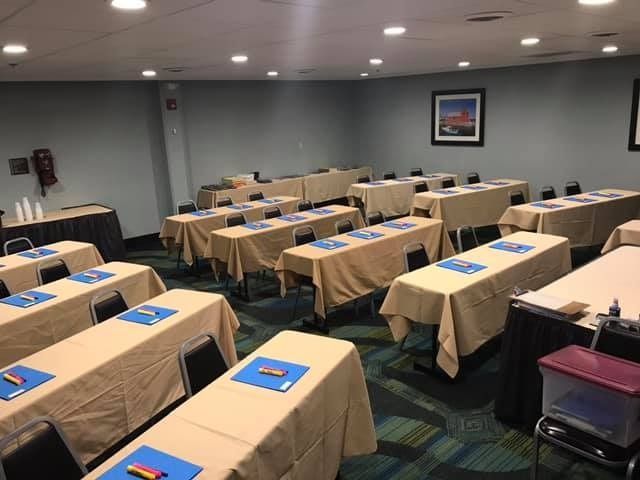 Classroom setup with tables covered in tan cloths, blue and yellow pens, and an event underway.