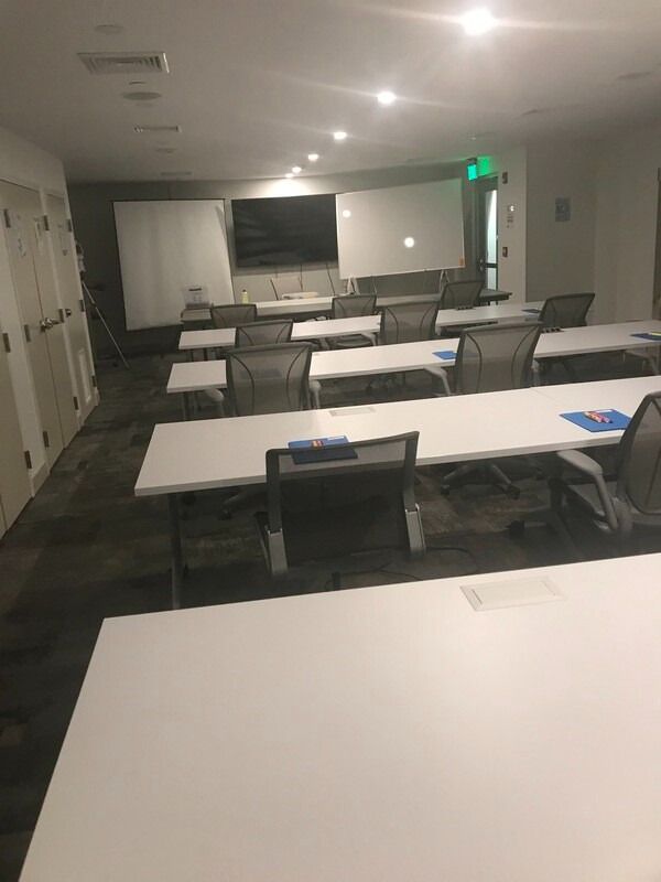 Empty classroom with white tables, chairs, whiteboards, and a TV on the wall.