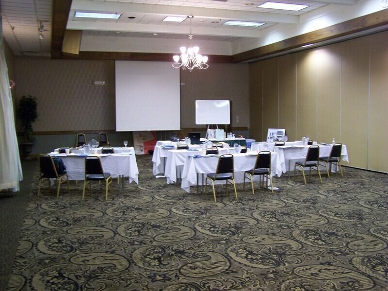 A meeting room with tables set for a presentation, white tablecloths, and black chairs, brown carpet.
