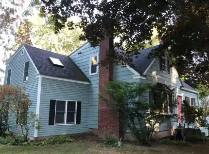 A large white house with a blue roof and black shutters is surrounded by trees.