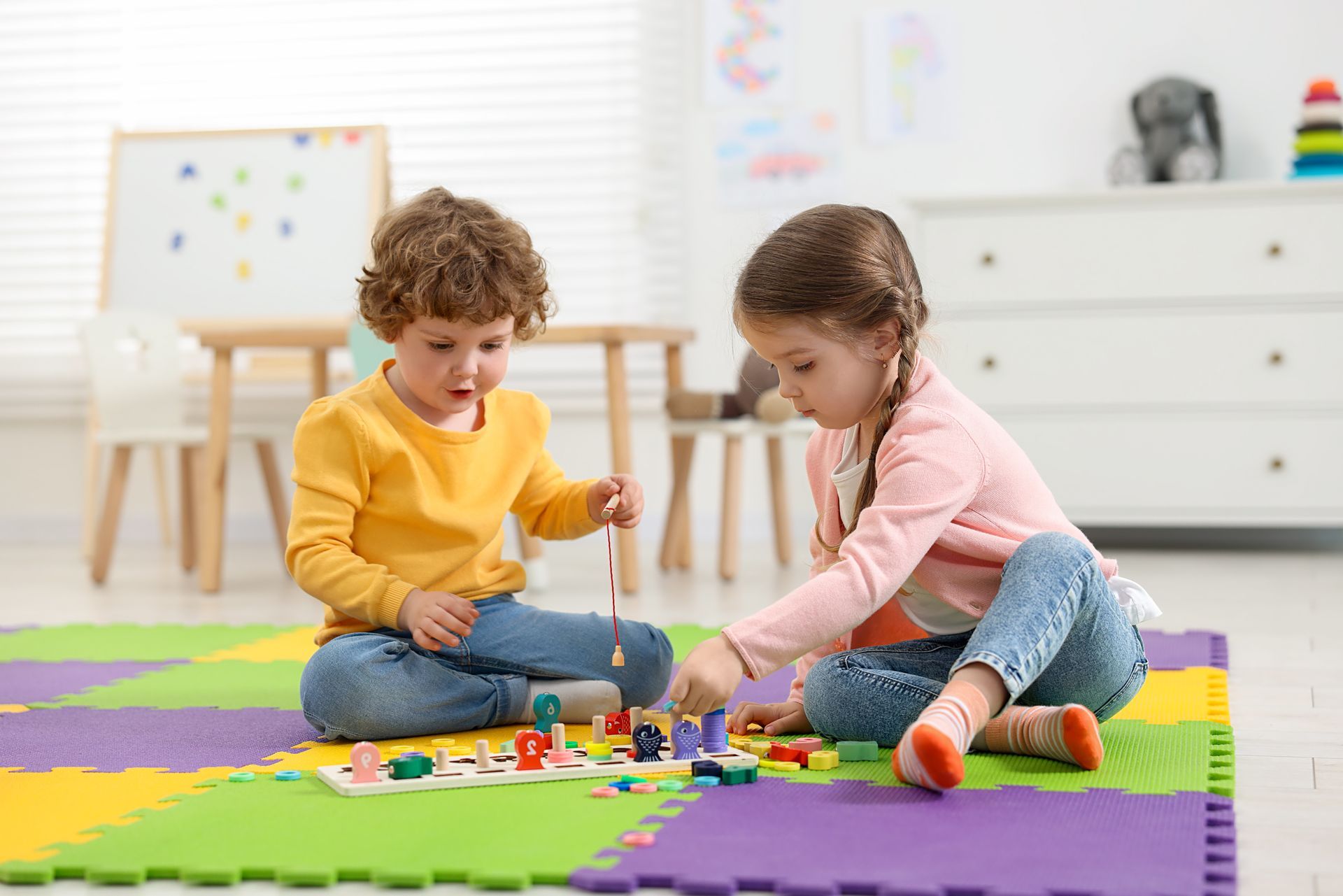 Two children playing a game with colorful wooden toys on a foam mat in a brightly lit room.