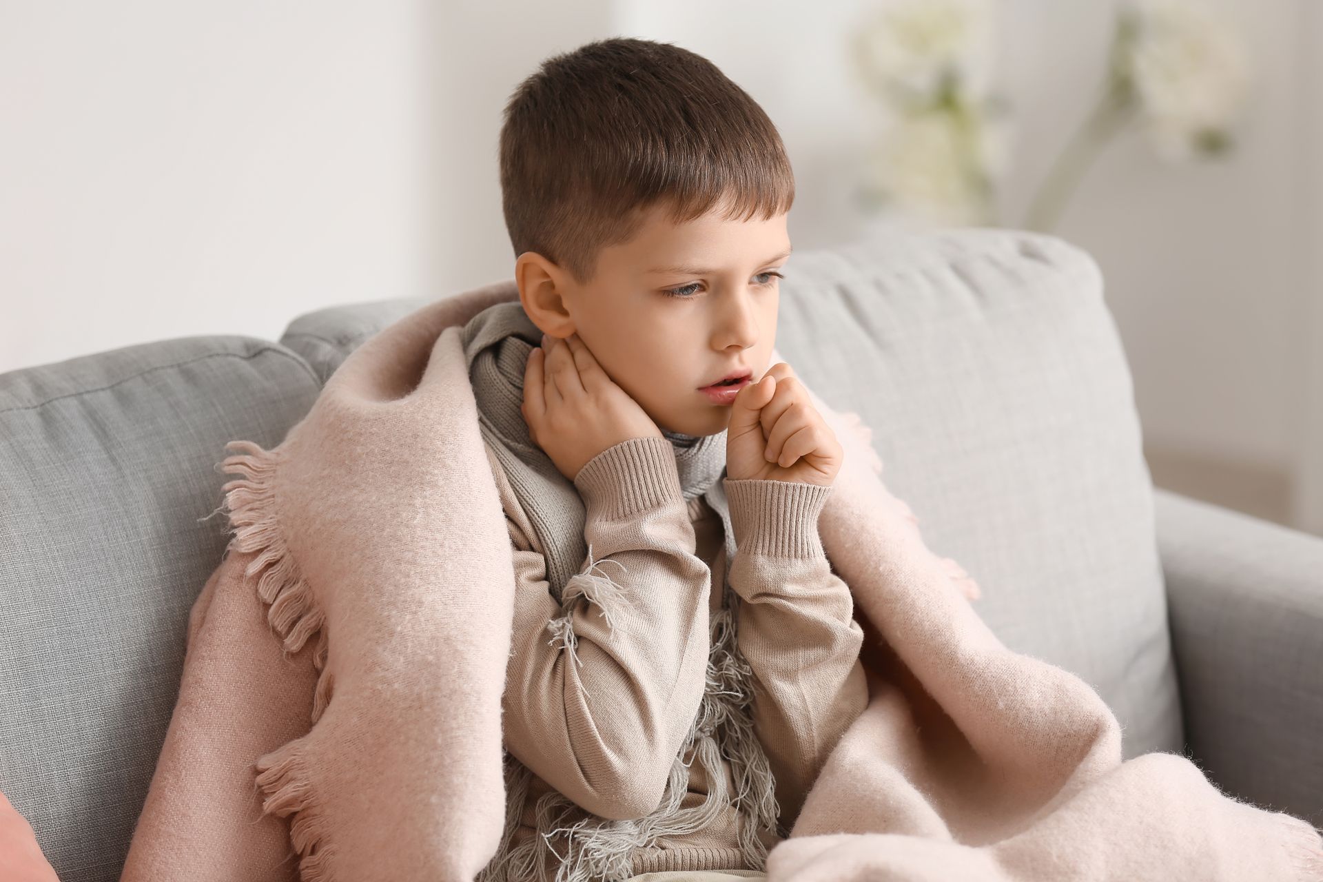 Boy wrapped in a blanket, coughing, sitting on a couch.