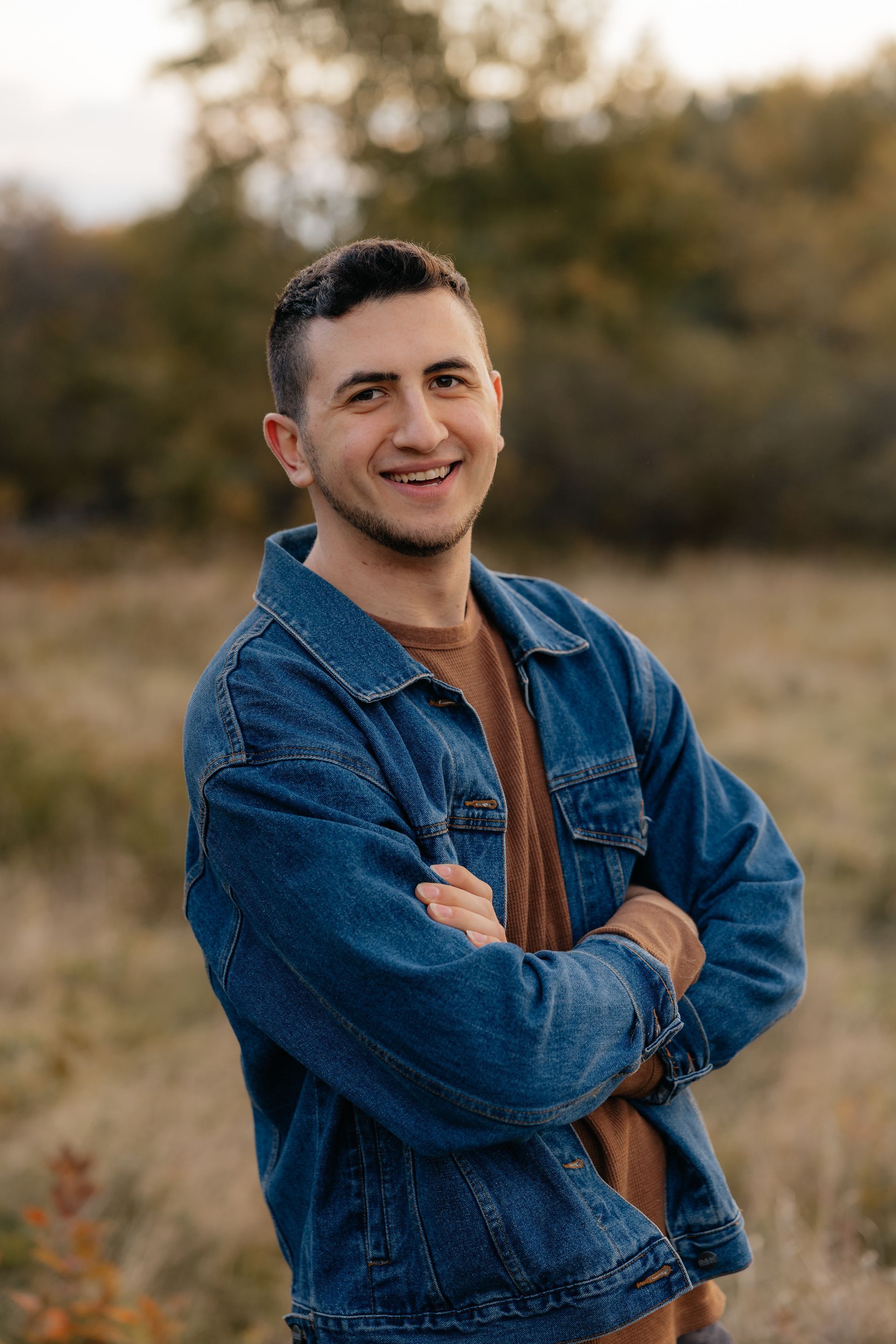 A man in a denim jacket is standing in a field with his arms crossed.