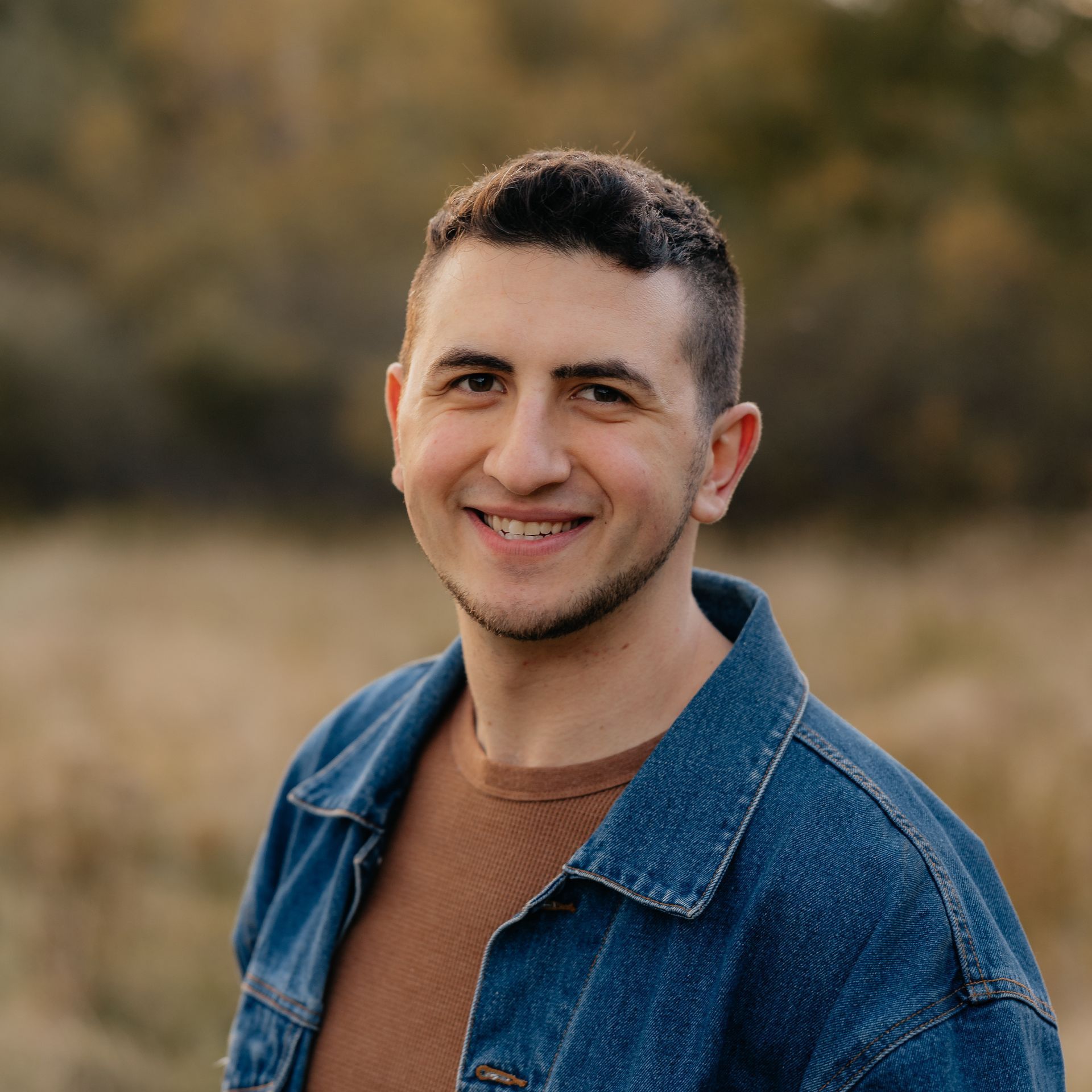 A man wearing a denim jacket and a brown shirt is smiling for the camera.