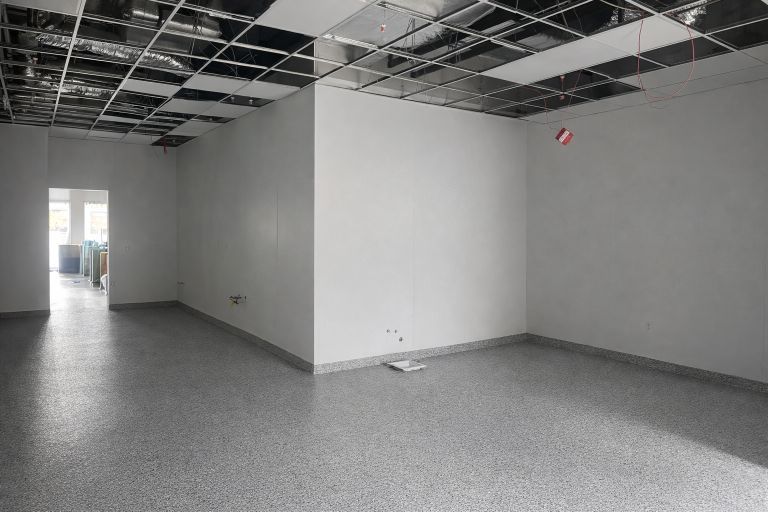 Empty room with exposed ceiling grid, white walls, and gray flooring.