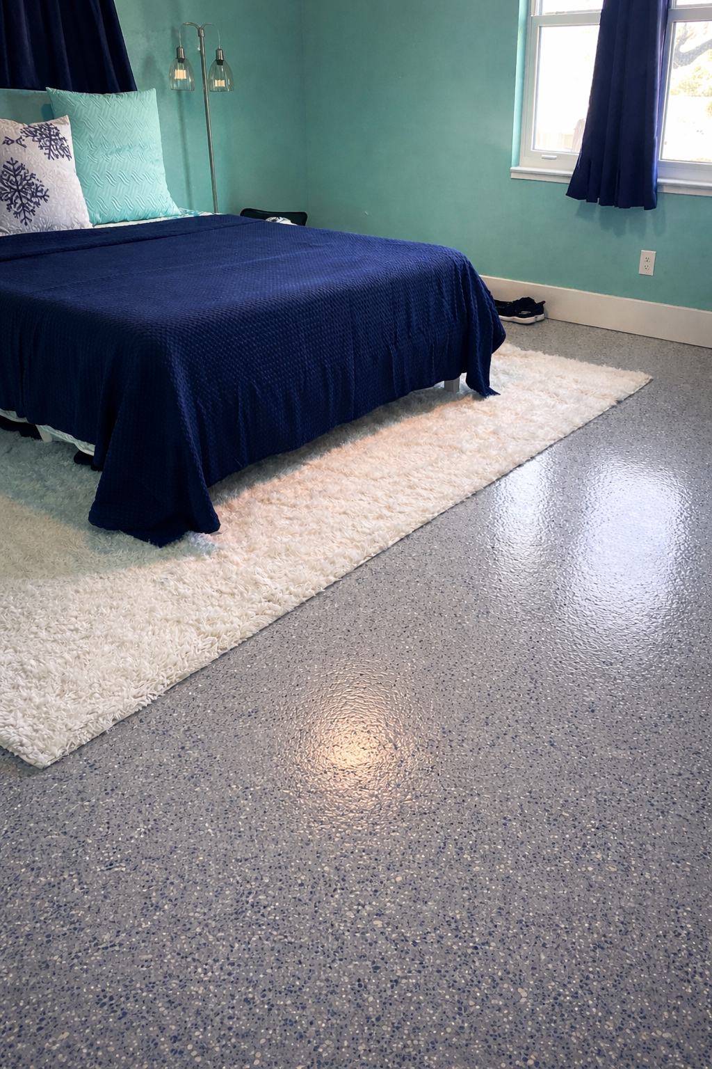 Bedroom with blue bedspread, turquoise wall, and speckled gray floor with a white rug.
