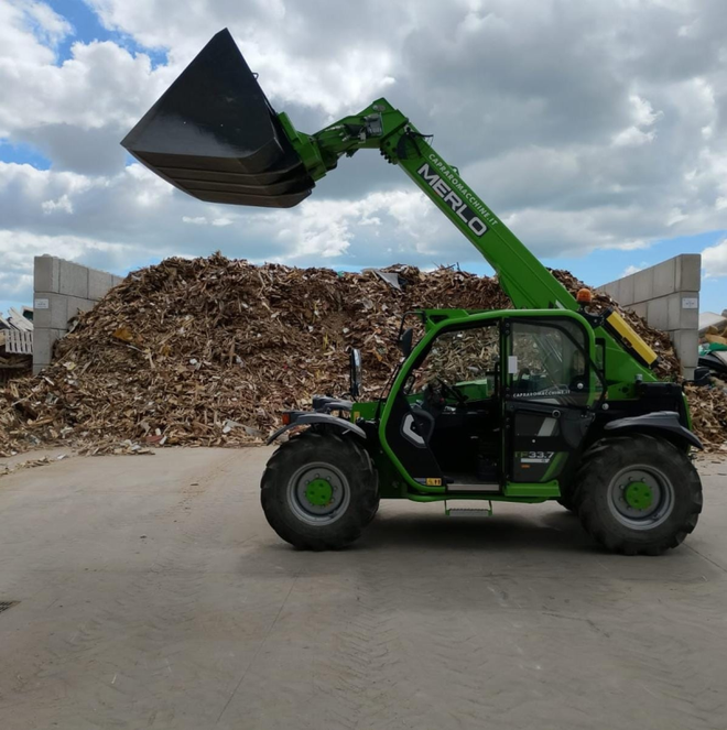 Un sollevatore telescopico Merlo verde, con la benna alzata, parcheggiato davanti a un grande cumulo di materiale di scarto su un piazzale in cemento.
