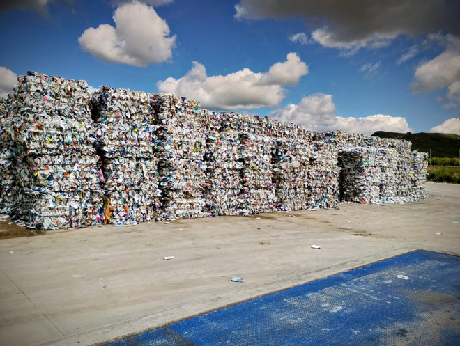 Grandi pile rettangolari di balle di plastica compressa e materiali riciclabili misti all'aperto, sotto un cielo azzurro con nuvole.