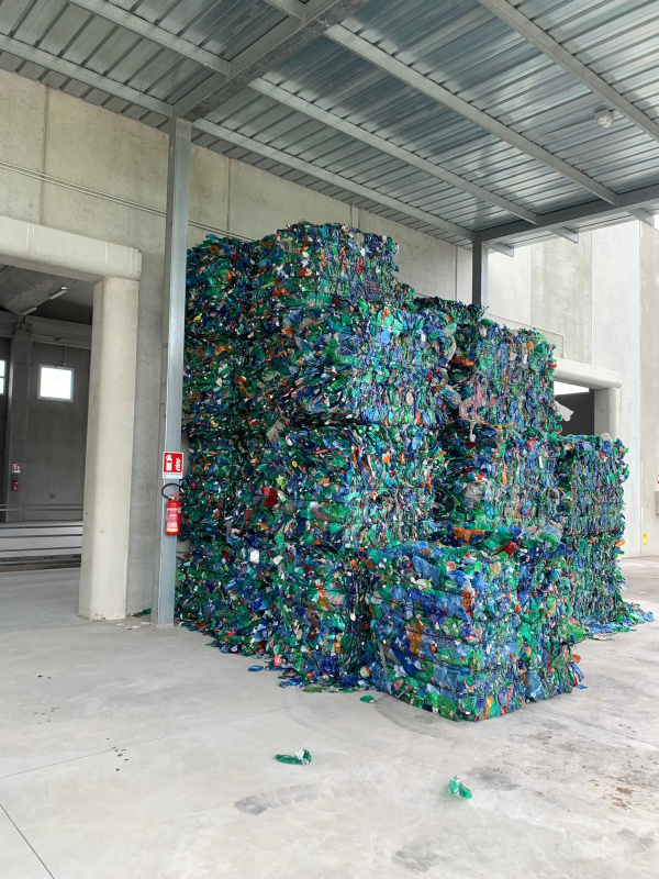 Un'ampia pila di bottiglie di plastica compresse e avvolte in balle, in un deposito sotto una tettoia metallica.