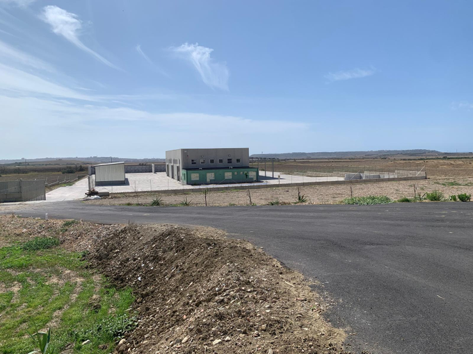 Un piccolo edificio di servizio con tetto piatto si erge in un paesaggio aperto, asciutto e recintato, sotto un cielo azzurro e limpido.