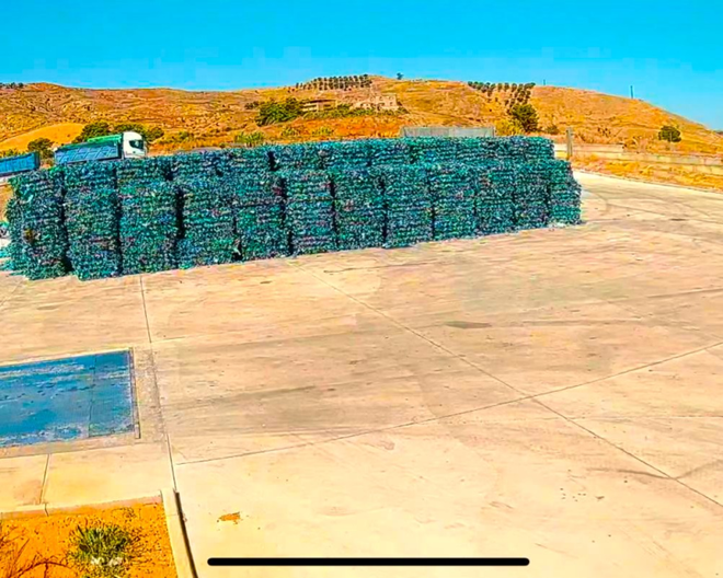 Una grande pila rettangolare di bottiglie di plastica blu compresse si erge su un piazzale industriale in cemento sotto un cielo azzurro e limpido.