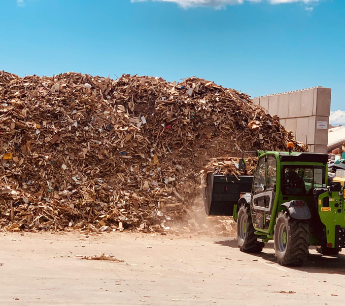 Un caricatore industriale verde sposta un grande cumulo di trucioli di legno in un'area di stoccaggio all'aperto, sotto un cielo azzurro e limpido.