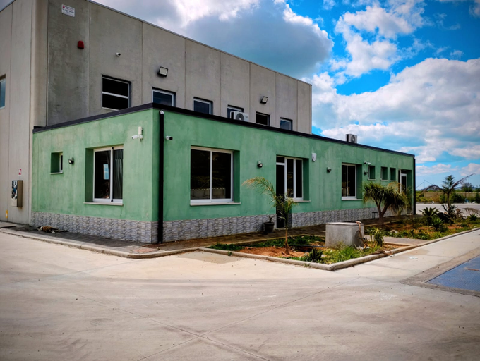 Un edificio verde a un solo piano, annesso a un magazzino in cemento più alto, sotto un cielo azzurro con nuvole bianche.